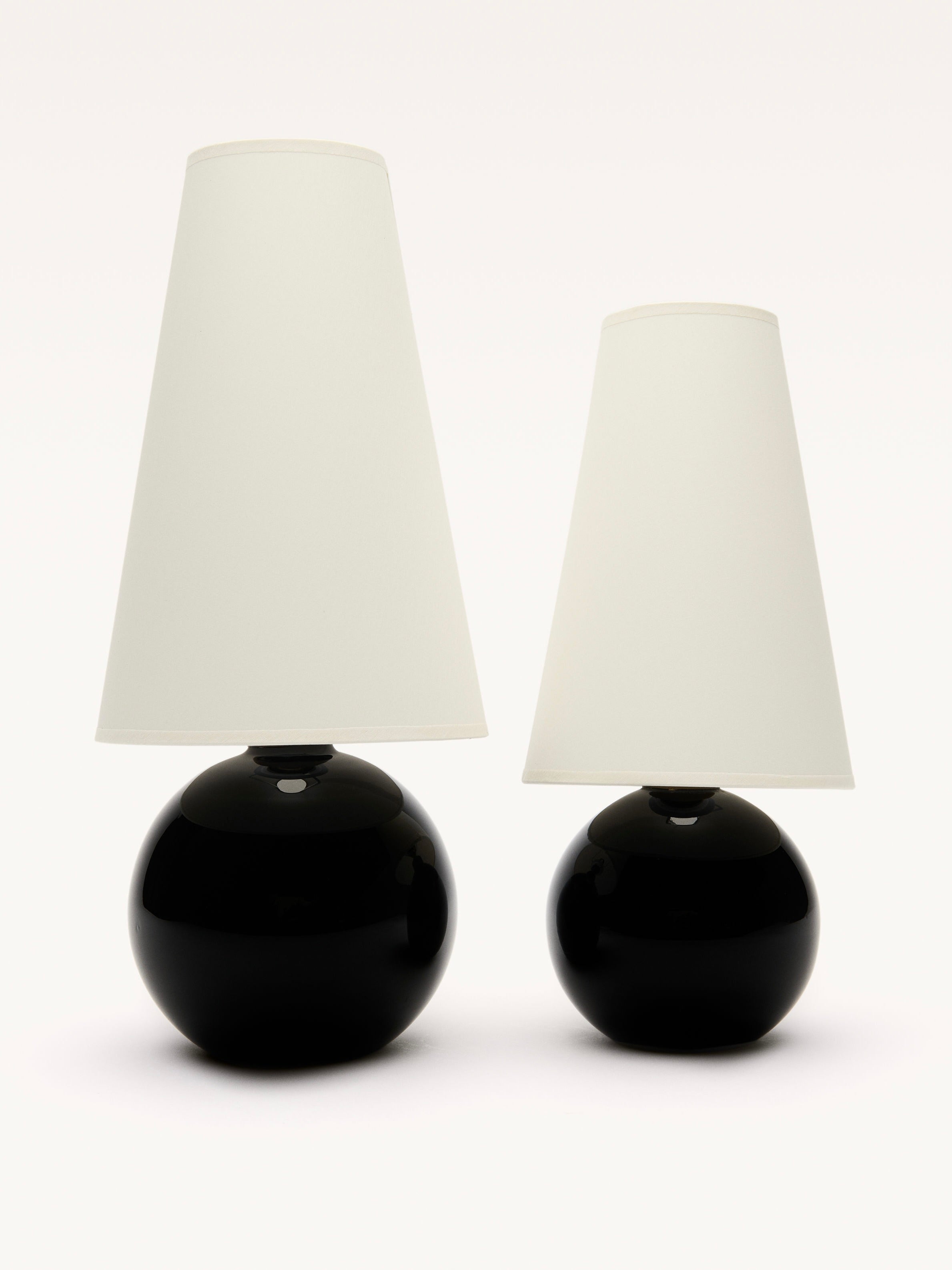 Two Galerie MLS lamps with black opalin glass bases and white cone-shaped shades stand against a white background. The taller left lamp evokes a Jacques Adnet style, enhanced by an elegant brass ring around its base.