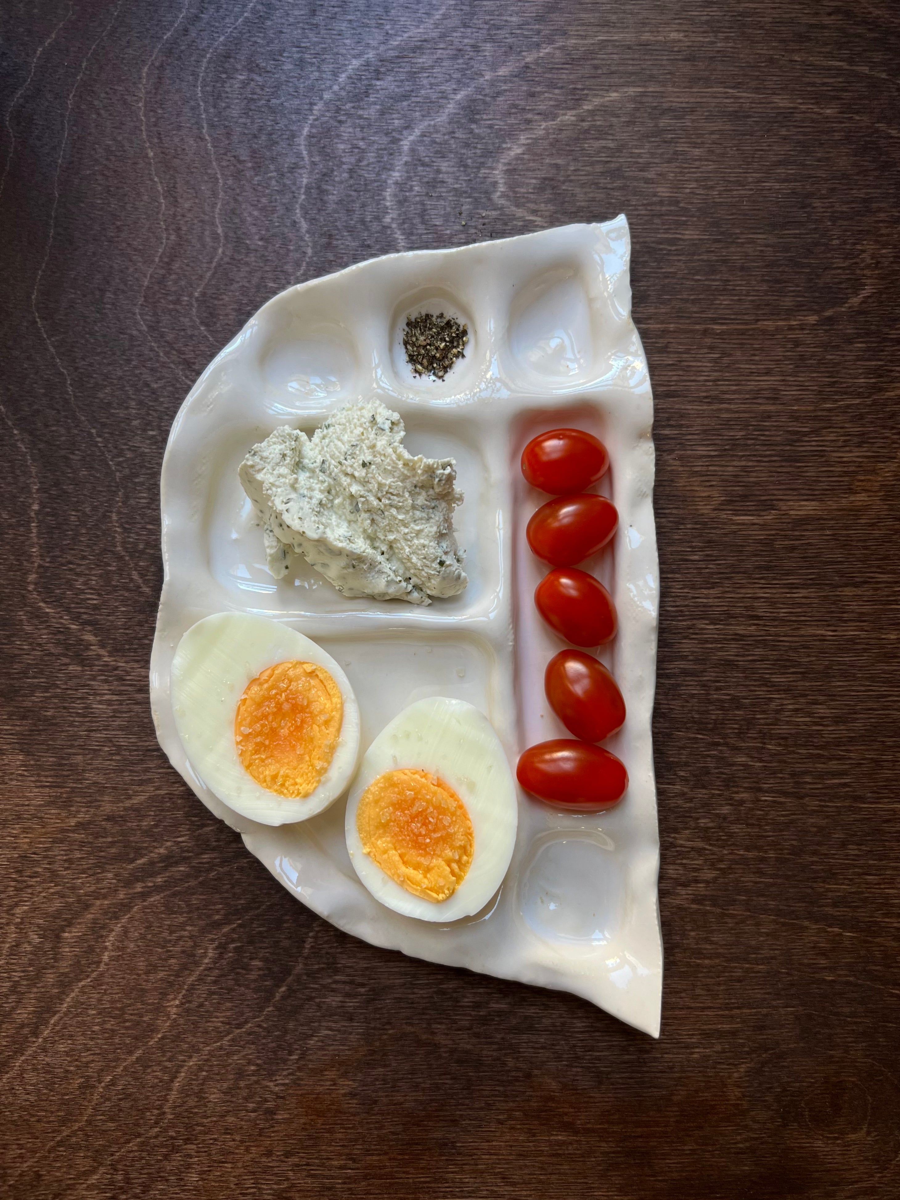 A uniquely shaped white clay Snack Plate with sections containing different foods: two halves of a boiled egg, a scoop of herbed cheese, a row of cherry tomatoes, and two small piles of spices by Somthings.