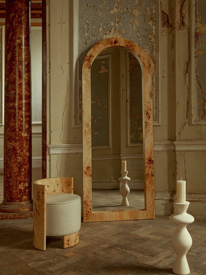 In a rustic room stands the Ransom & Dunn Arc Burl Wood Floor Mirror, its arched design framed in wood, alongside a stylish wooden chair with a white cushion. A sculptural candle holder decorates the ash burl wood floor while the walls' peeling paint contributes to a vintage atmosphere with neoclassical architectural hints.