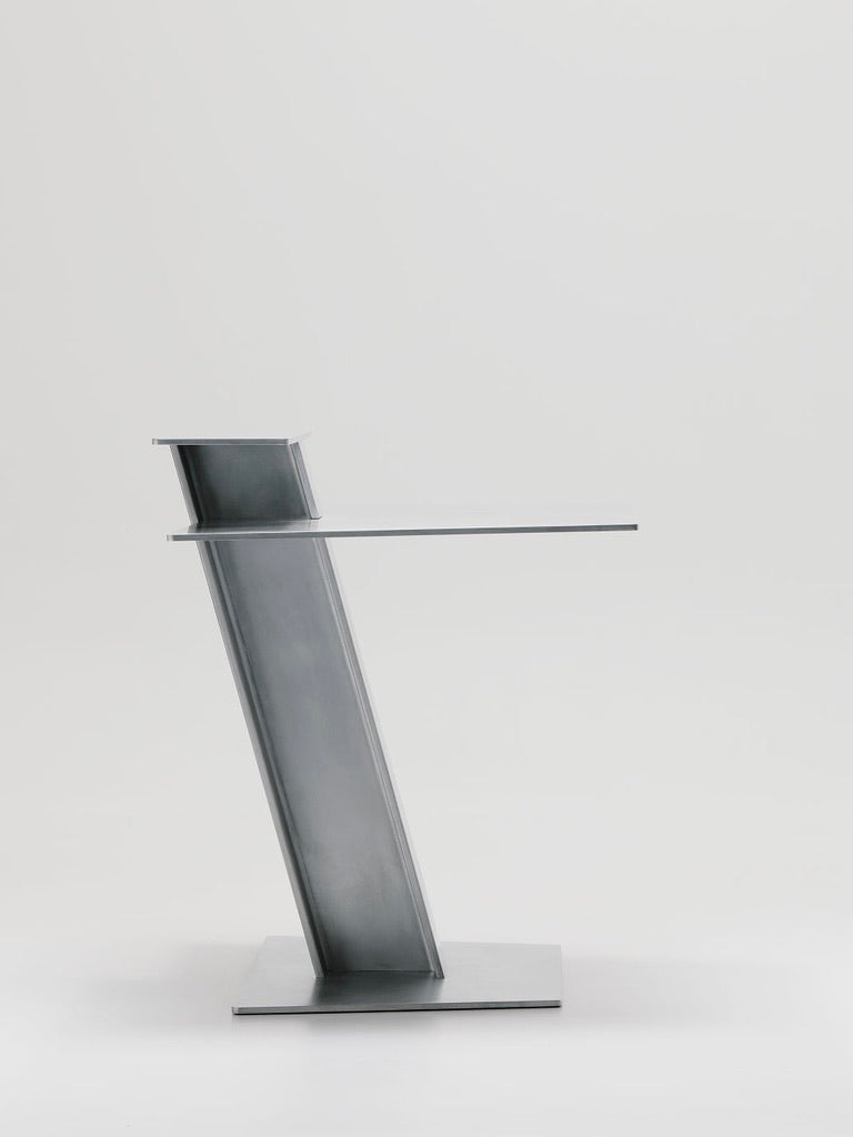 The Beam Desk by Marquel Williams is a minimalist gray lectern with an angled design, featuring a sleek metal finish and unique slanted structure against a white background, enhancing its modern and contemporary aesthetic.