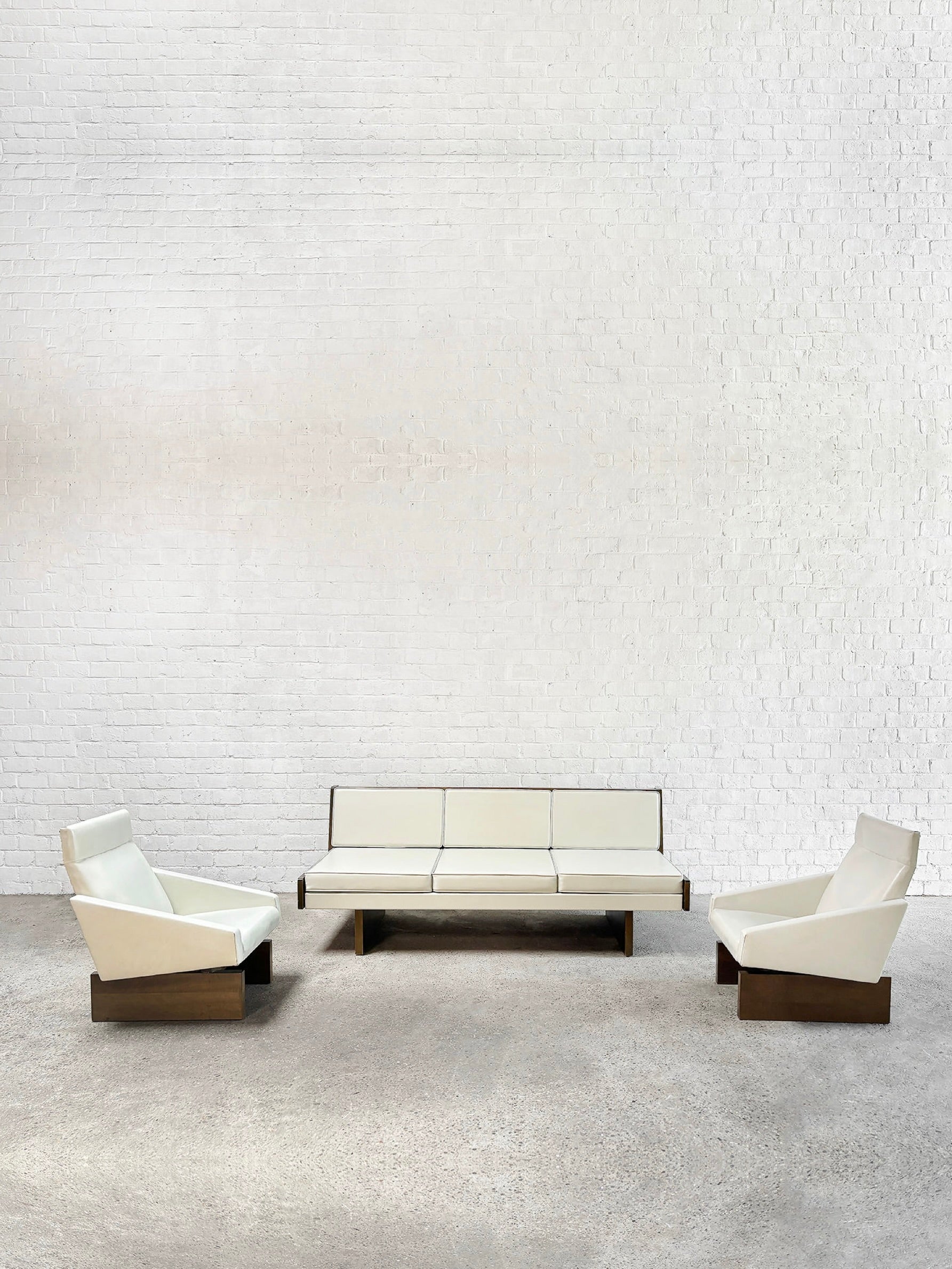 Create a stylish minimalist seating area with Introverso's French Modernist Sofa Set in white eco-leather. This 1950s-inspired set includes a three-seater sofa and two matching armchairs, all with wooden bases, perfect against a white brick wall and grey concrete floor.