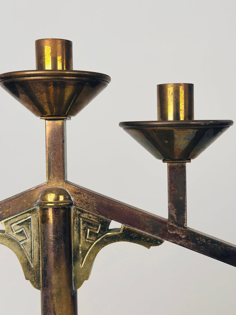 A close-up of a Figuresdesign Modernist Candleholder reveals its two candle sockets placed at different heights. The metal, in good condition, features a slightly tarnished, aged appearance with a nice patina. The design includes some ornamental detailing, set against a plain, light gray background.
