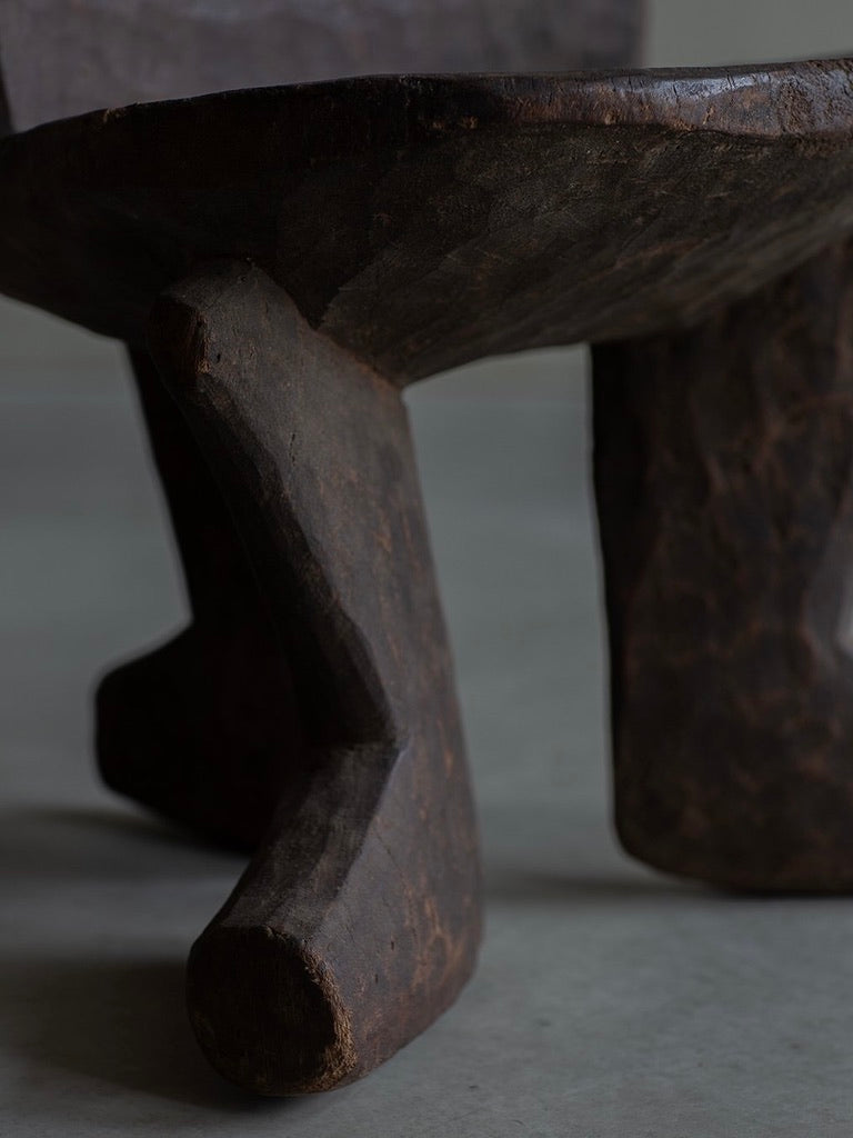 A close-up of Bicci de' Medici's African Stool highlights its rustic, three-legged wooden design, dark textured wood reflecting natural light. The primitive style underscores African artisans' craftsmanship, with a blurred background emphasizing its cultural heritage and rough surface.