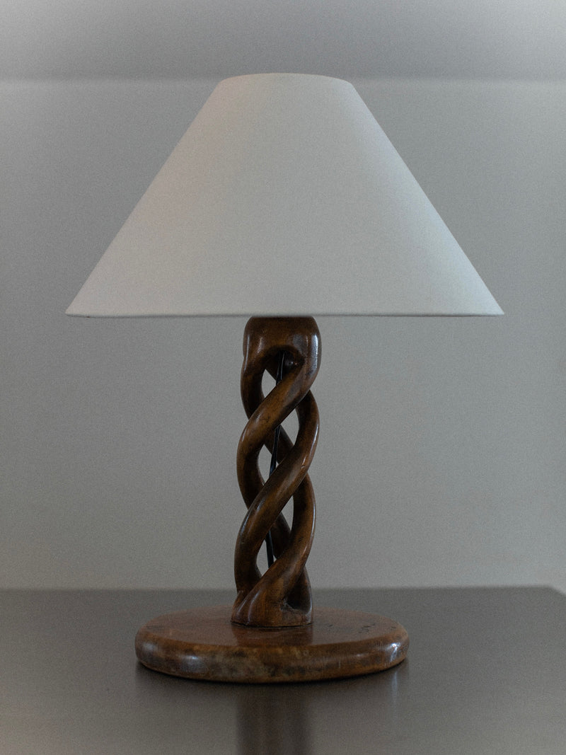 The Early-Century Lamp by Out For Lunch is a handcrafted piece with a white conical shade and a twisted wooden base adorned with an intricate swirling design, resting on a smooth, dark surface against a neutral backdrop.