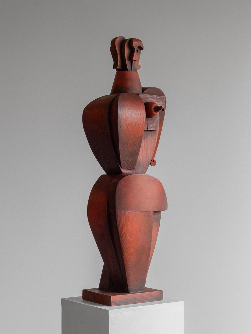 The Vintage Cubist Sculpture II by Atelier Boulogne from Spigel is a tall wood piece with a smooth, reddish-brown finish. It showcases geometric shapes forming a stylized female figure with broad shoulders, a tapered body, and an abstract head on a white pedestal against a neutral background.