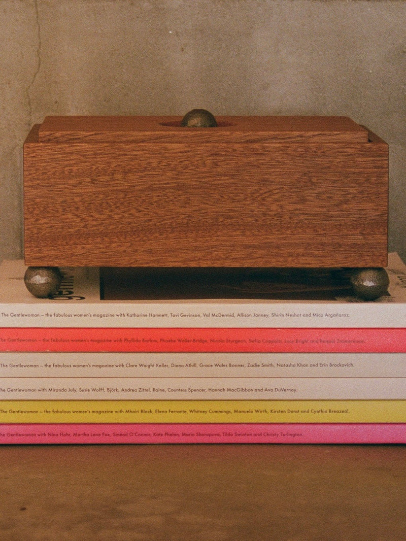 A stack of magazines with colorful spines is topped by The Phone Box from MUHLY, a handmade wooden box from Philadelphia featuring a smooth finish. The magazines are neatly arranged, and the box’s small round knobs slightly elevate it above the stack, promoting phone-free living in your cozy reading nook.
