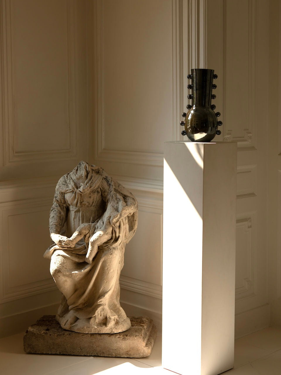 A headless stone sculpture rests on the floor beside a white pedestal in a softly lit room. Atop the pedestal is the Vase Solstice II by Landy Rakoto Ceramics, featuring a shiny, textured surface with a striking metallic effect finish. Sunlight filters through, casting shadows on the wall.