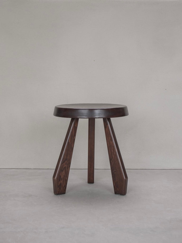 The Charlotte Perriand Sandoz Stool by Bicci de' Medici, featuring a circular seat and three angled legs, stands against a plain light gray background. The minimalist design emphasizes the mahogany wood grain and craftsmanship.