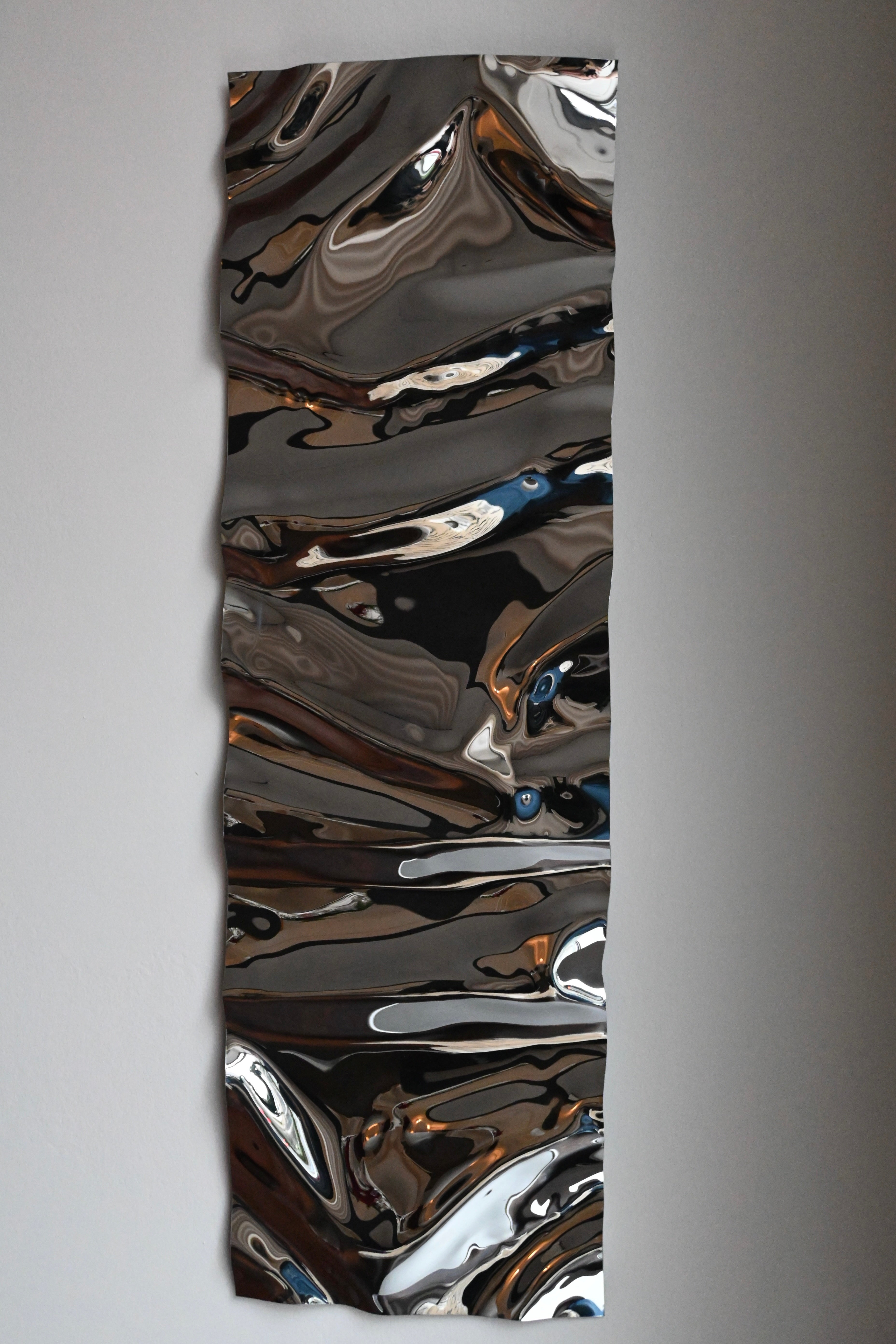 The vertical Psychedelic Mirror Long by Caia Leifsdotter features wavy, metallic reflective surfaces that resemble a handmade sculptural mirror. Its polished finish creates fluid, distorted reflections, showcasing unique artistry. It's mounted on a plain light gray wall.