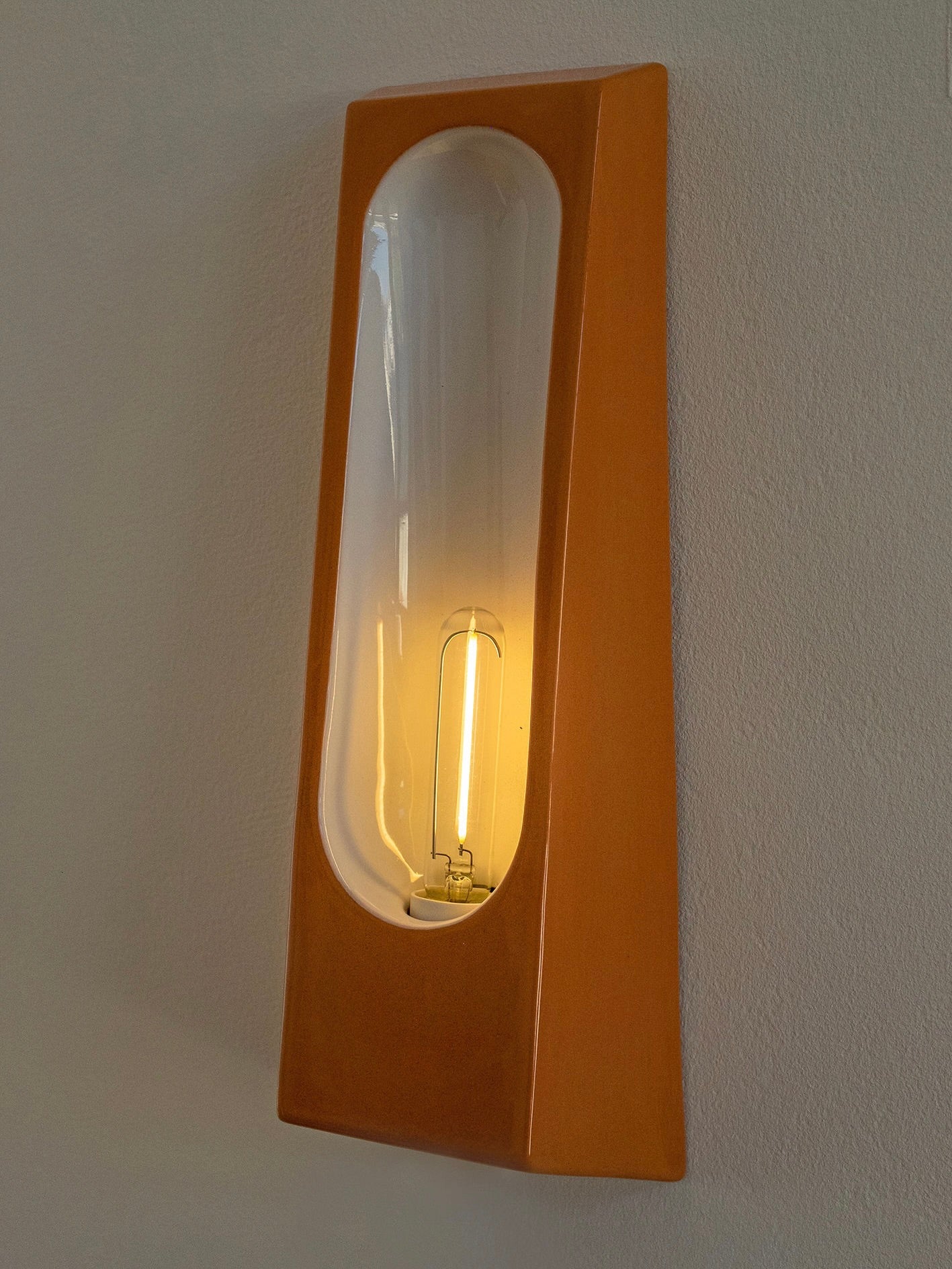 The Alcove Terra B Glossy by Violaine d'Harcourt is a modern wall light with a sleek, rectangular design in warm wood tones and clear elongated glass. It reveals a glowing bulb casting soft illumination, incorporating glazed ceramic for an elegant finish.