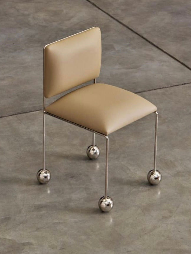 Introducing Panorammma's 'Ball Foot' Chair: a chic design featuring a beige vegan textile seat and backrest. Supported by thin metal legs with shiny, large spheres, it rests elegantly on a gray concrete floor, weaving together style and sustainability.