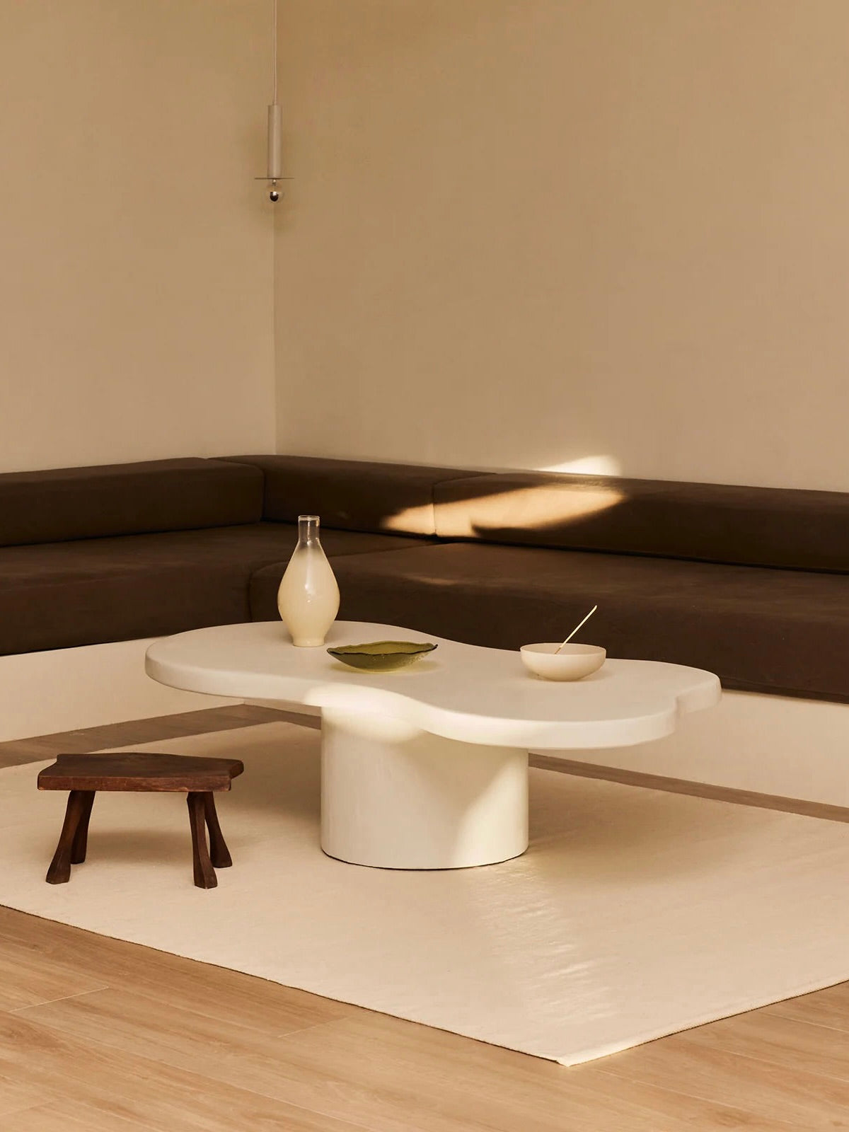 A minimalist living room showcases a beige corner sofa and the artisan-crafted MarlotBaus "Pesto" Coffee Table, adorned with a white vase and bowl. A small wooden stool rests on a light area rug atop microcement flooring, while soft lighting enhances the neutral color palette.