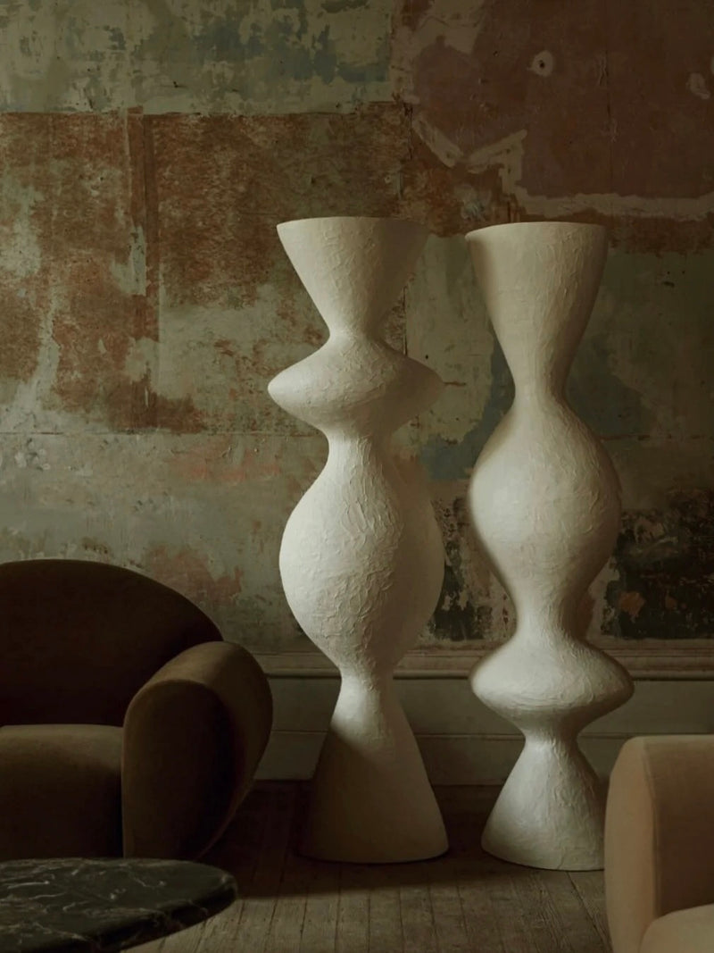 Two tall, abstract white sculptures with textured surfaces stand side by side in a dimly lit room. Handmade in Portugal, these pieces add elegance against the aged, distressed wall. On the left, a curved brown armchair accompanies an eco-resin up-lighter known as the "Venus II" Floor Lamp by Ransom & Dunn.