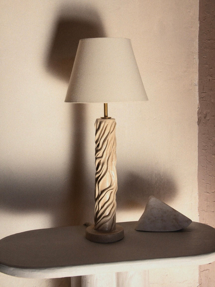 The "Waves" Table Lamp by Lorea Palacios, featuring a textured ceramic base and white shade, sits on a small table next to a triangular marble-like ornament. This contemporary interior is warmly lit, casting soft shadows on the wall behind.