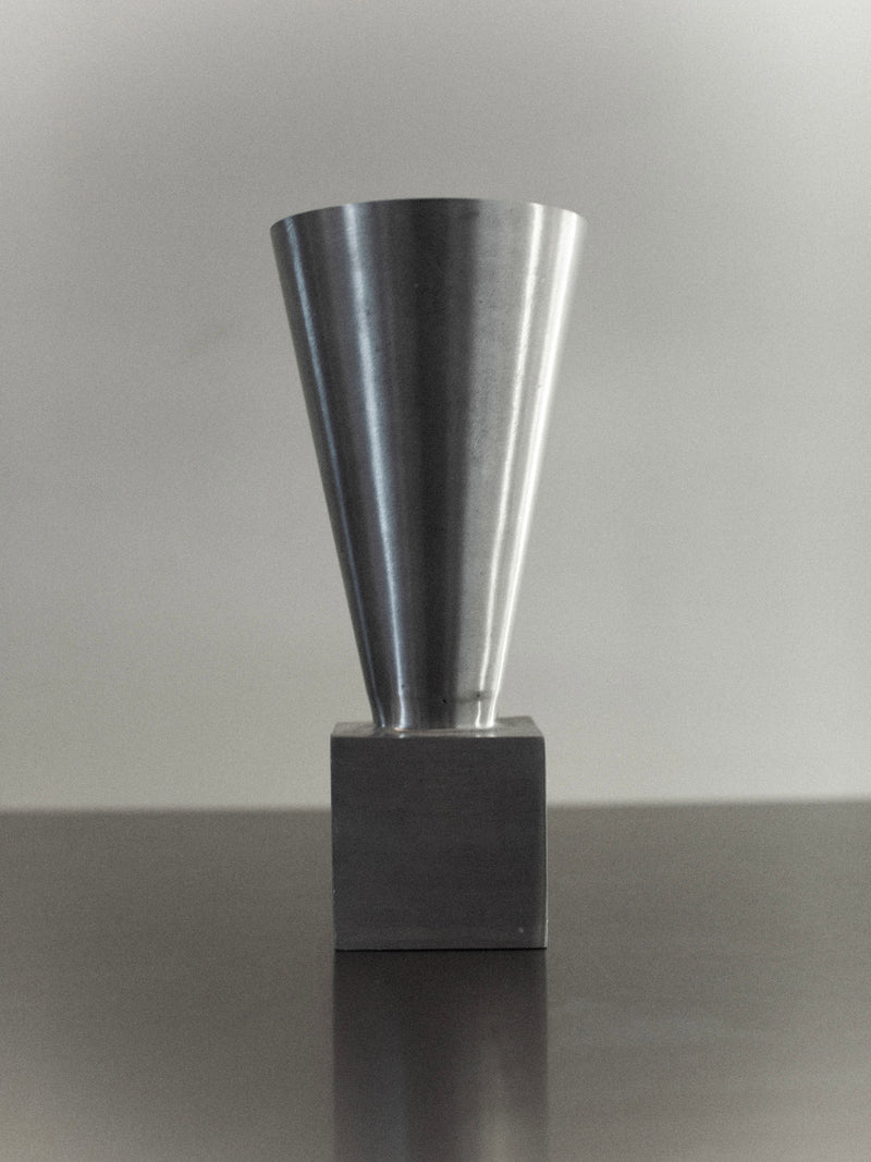 The 70's Aluminum Cast Vase by Out For Lunch showcases a sleek minimalist design with its conical top and square base, crafted from brushed aluminum. Set against a neutral background, this modern piece emphasizes clean lines and a reflective surface.