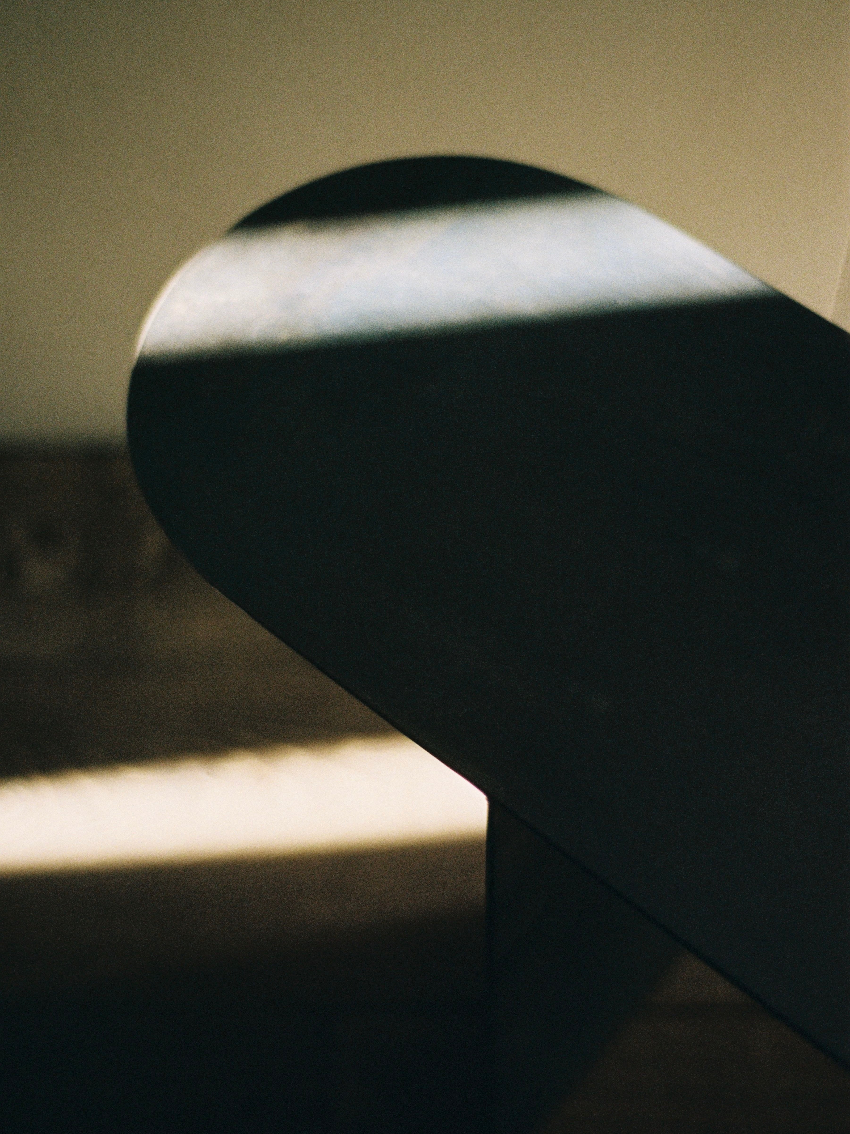 Sunlight casts a delicate shadow on the curved, dark surface of The Altar by COSEINCORSO, crafting an abstract and serene atmosphere. The interplay of light and shadow accentuates the gentle curves and texture of the material, reminiscent of limited edition prayer kneelers.