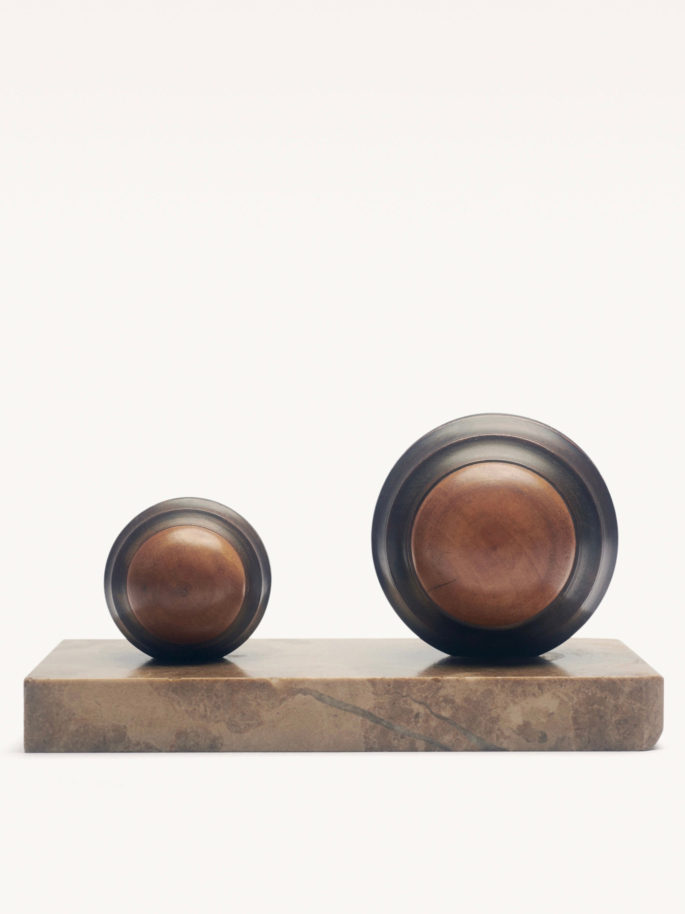 The Galerie MLS Pair of Art Deco Paper Press features two modernist wooden spheres with dark metallic rims on a rectangular marble base. The larger sphere is on the right, smaller on the left, all set against a plain white background to highlight their minimalist allure.