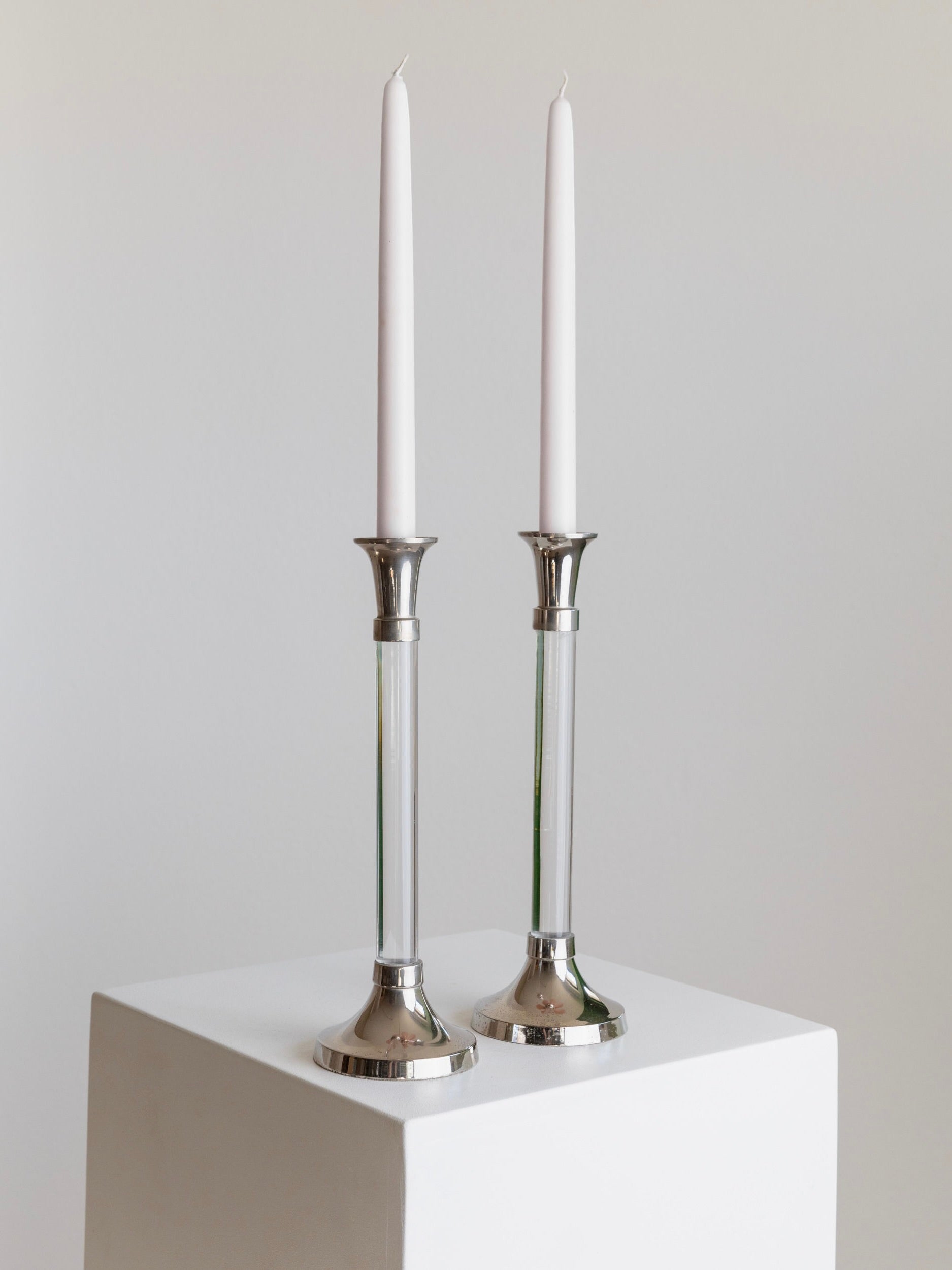 The Set of 2 Candleholders by Bottega Jacobs features two elegant white taper candles standing tall in vintage-style holders crafted from chrome-plated metal and acrylic glass. Placed on a white cube against a plain light background, they create a minimalist and modern aesthetic.