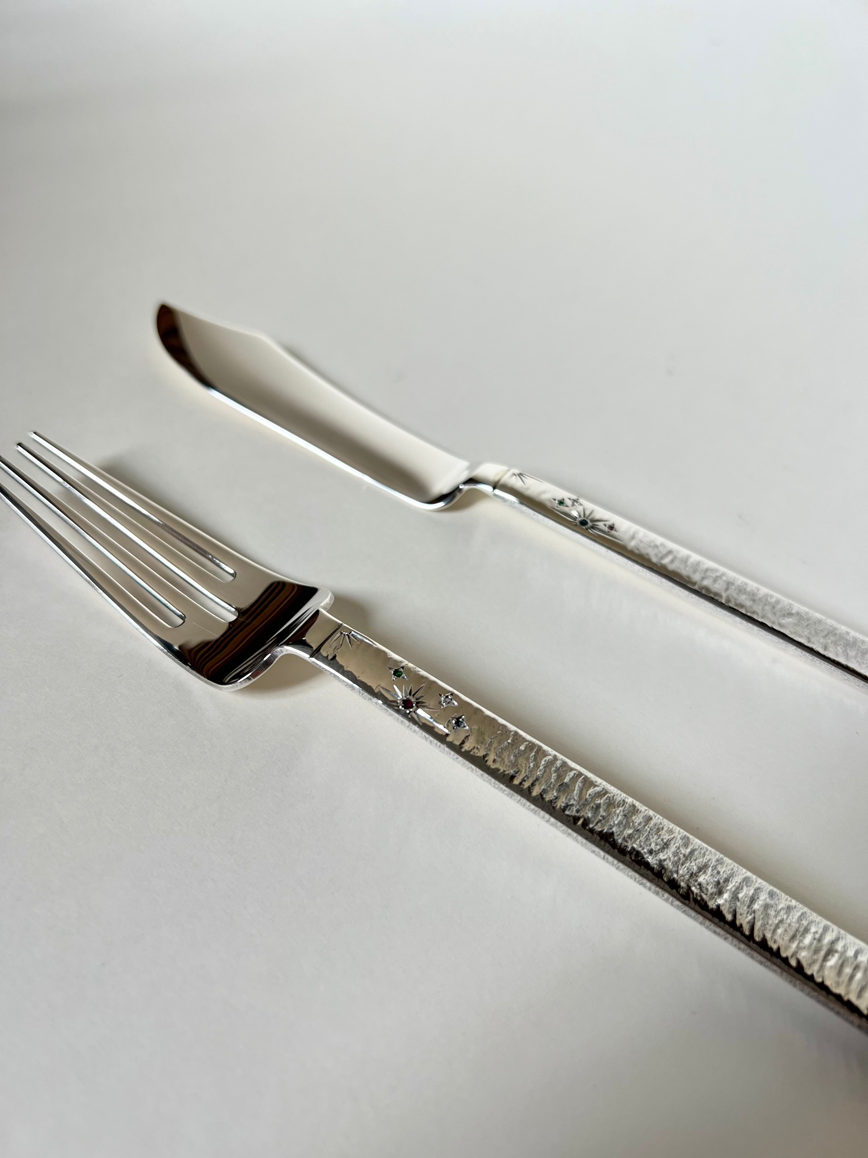 Sentence with replaced product and brand name: A 2 Piece Silver Cutlery set from Tavares 1922, with ornate handles, crafted from 925 Silver, are placed parallel on a white surface, reflecting light on their polished surfaces.