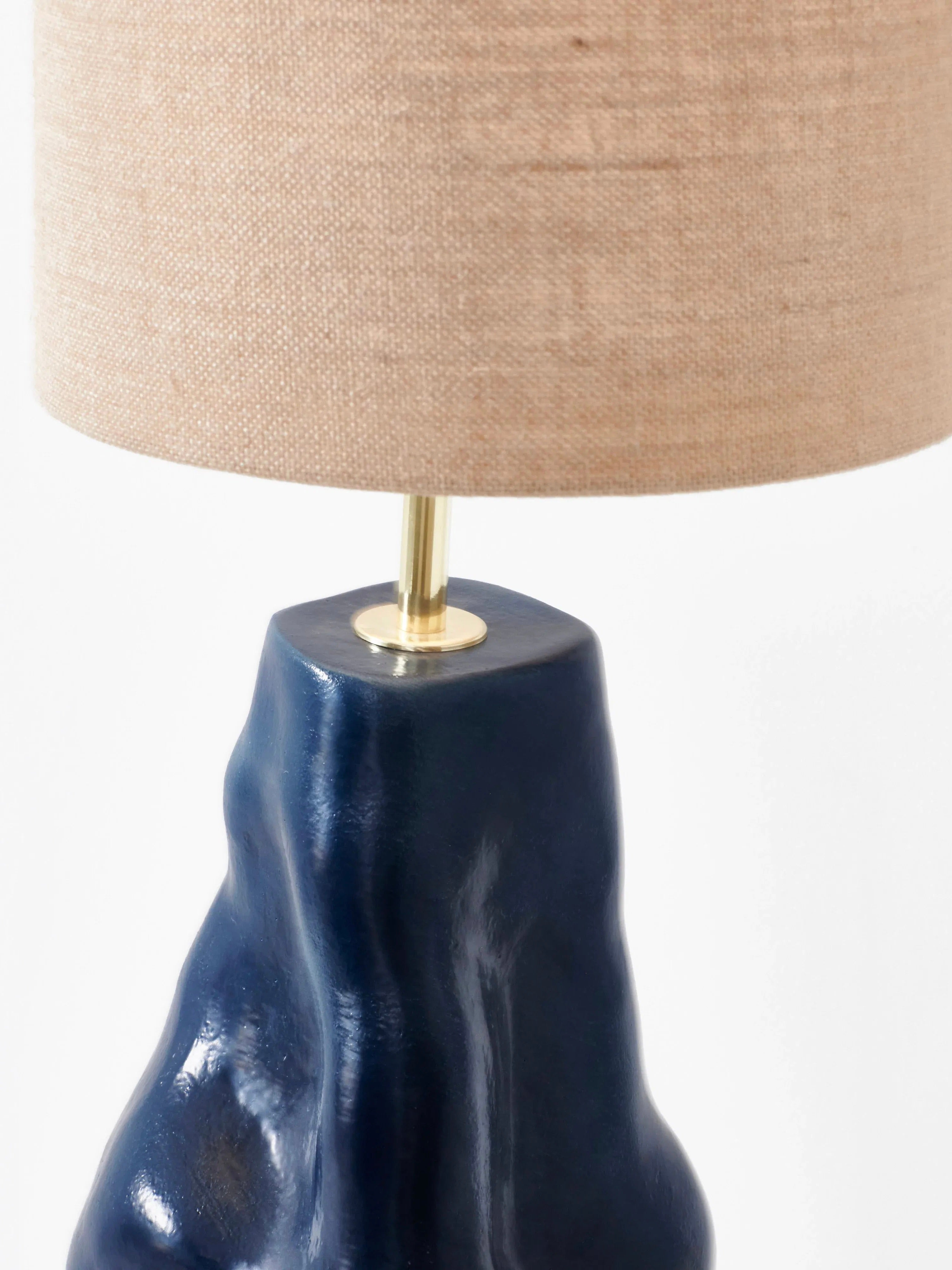 A close-up of the "Menina" Floor Lamp in Blue by Project 213A, showcasing a beige fabric lampshade and a shiny, textured dark blue ceramic base. Handmade in Northern Portugal, this artisanal floor light features an organic, irregular shape that lends it a modern and artistic appearance.