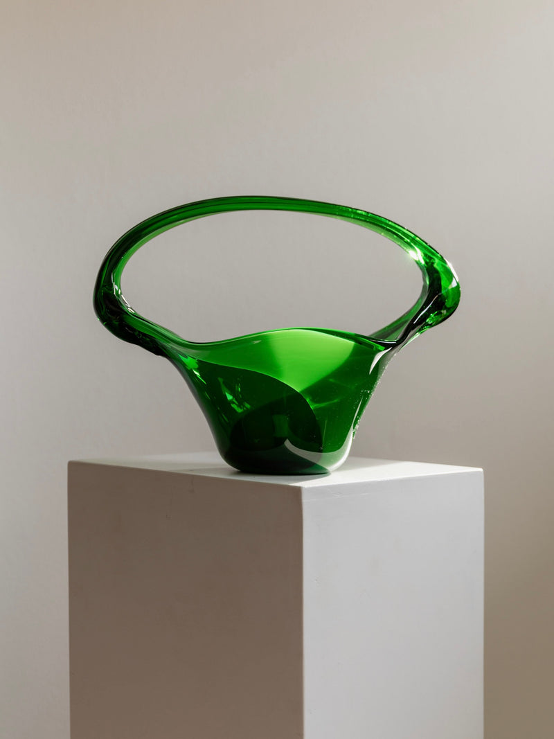 The Bottega Jacobs Murano Glass Handle Bowl 70s, in translucent green, flaunts an open, flowing design with a looped handle. Resting on a white pedestal against a neutral backdrop, it embodies artistic and abstract elegance reminiscent of Sommerso Murano craftsmanship.