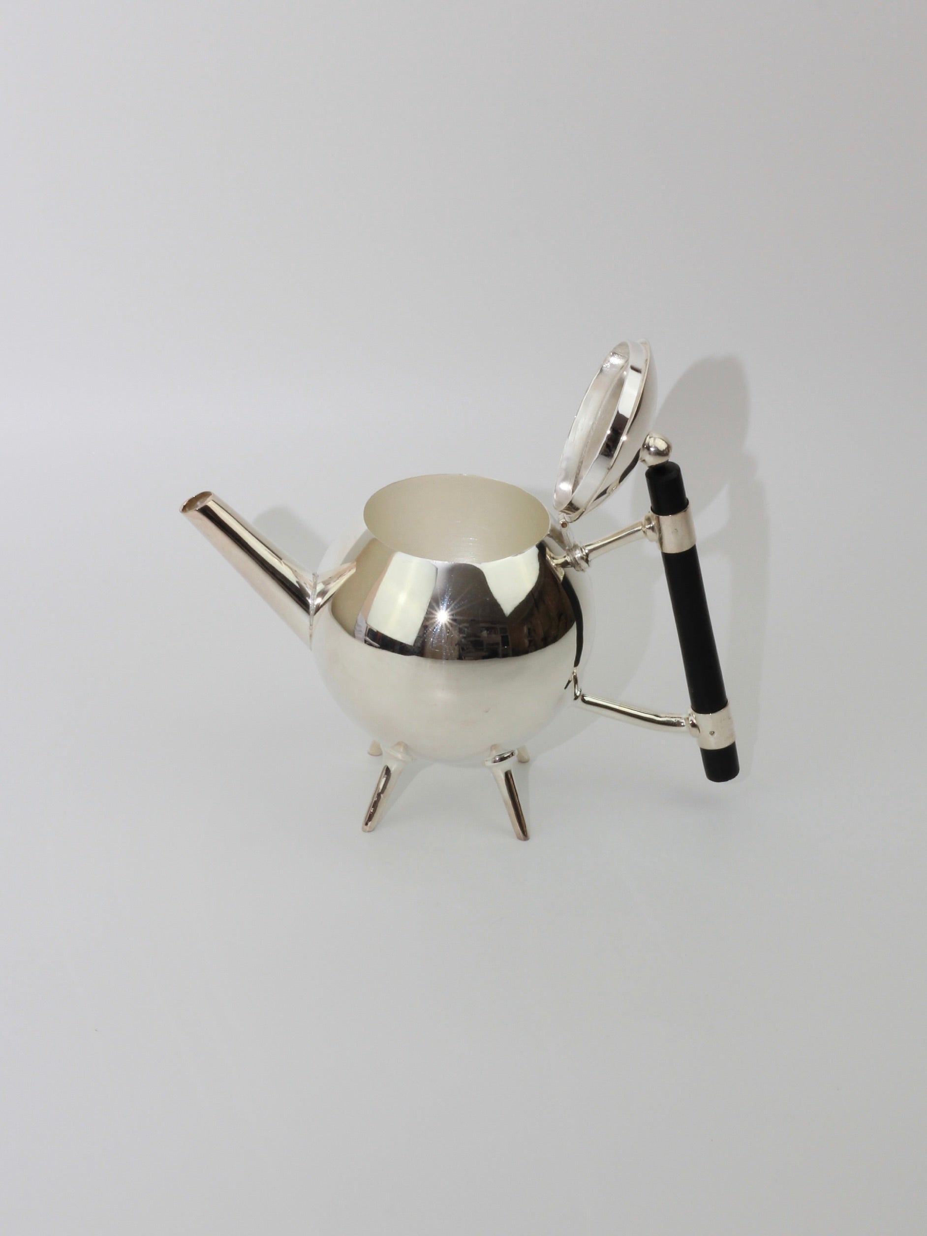 Titled "Christopher Dresser's Tea Pot," this stylish and contemporary stainless steel teapot from Boga Avante Shop features a spherical body, a slim spout, and an ebonized wood handle. The silver-plated teapot is reminiscent of Dr. Christopher Dresser's iconic designs, elevated on three short legs with its hinged lid open, elegantly placed against a plain white background.