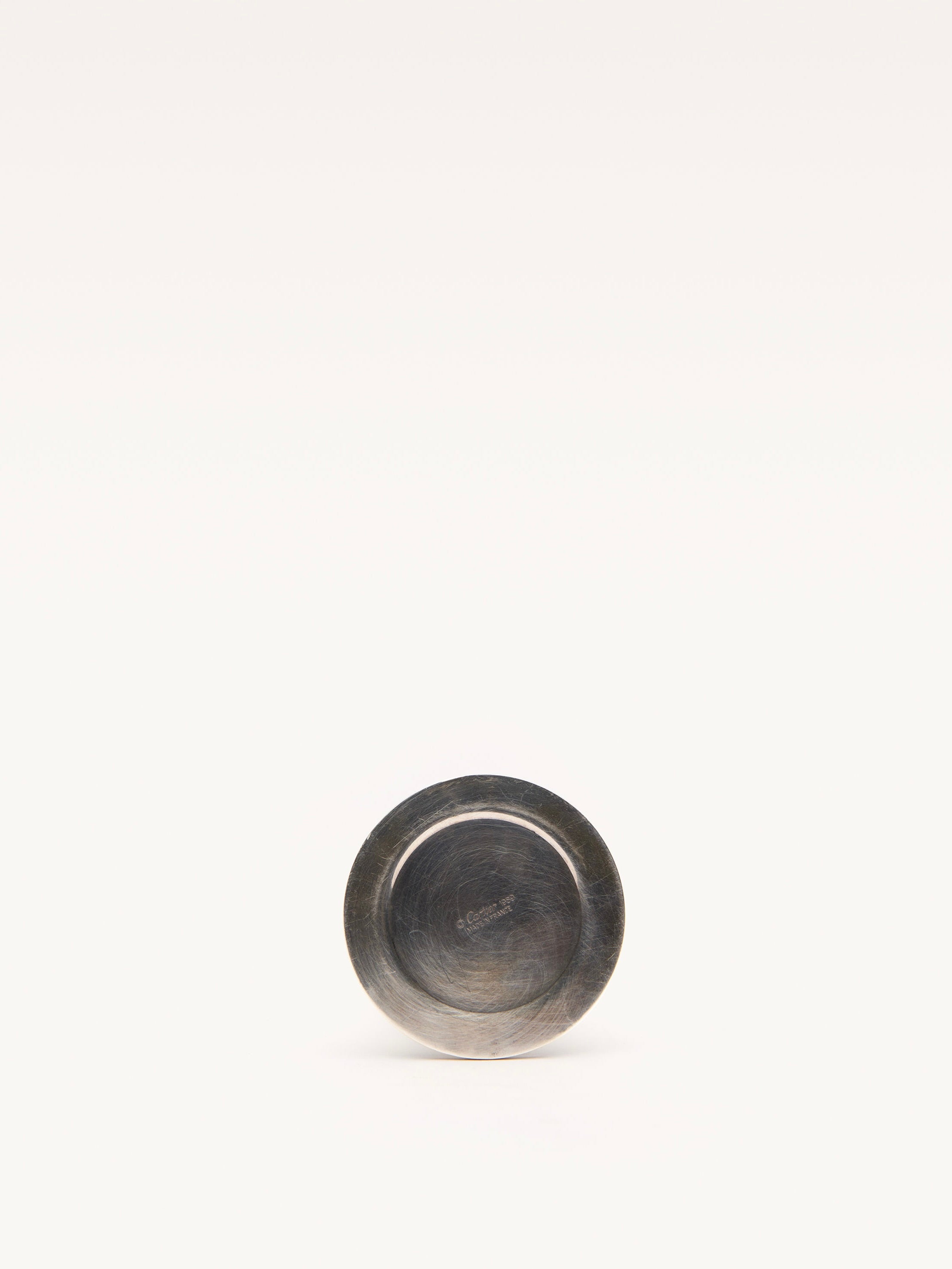 A small, round, silver-plated metal object with engraved text resembles an elegant Galerie MLS Cartier Candle Holder, positioned upright against a plain, light background.