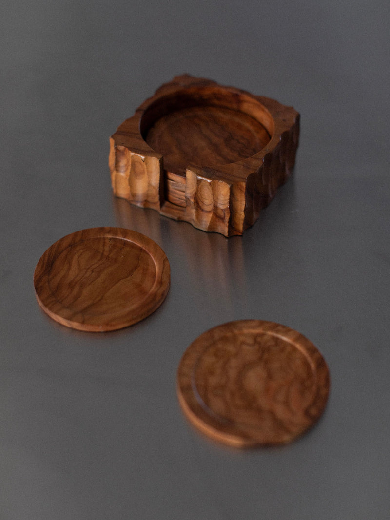 A photo featuring Out For Lunch’s Handcarved Coasters shows two round wooden coasters with detailed handcarvings, alongside a square wooden coaster holder with rounded edges, all arranged on a smooth gray surface. The natural elegance of the coasters and holder is highlighted by their visible wood grain and polished finish.