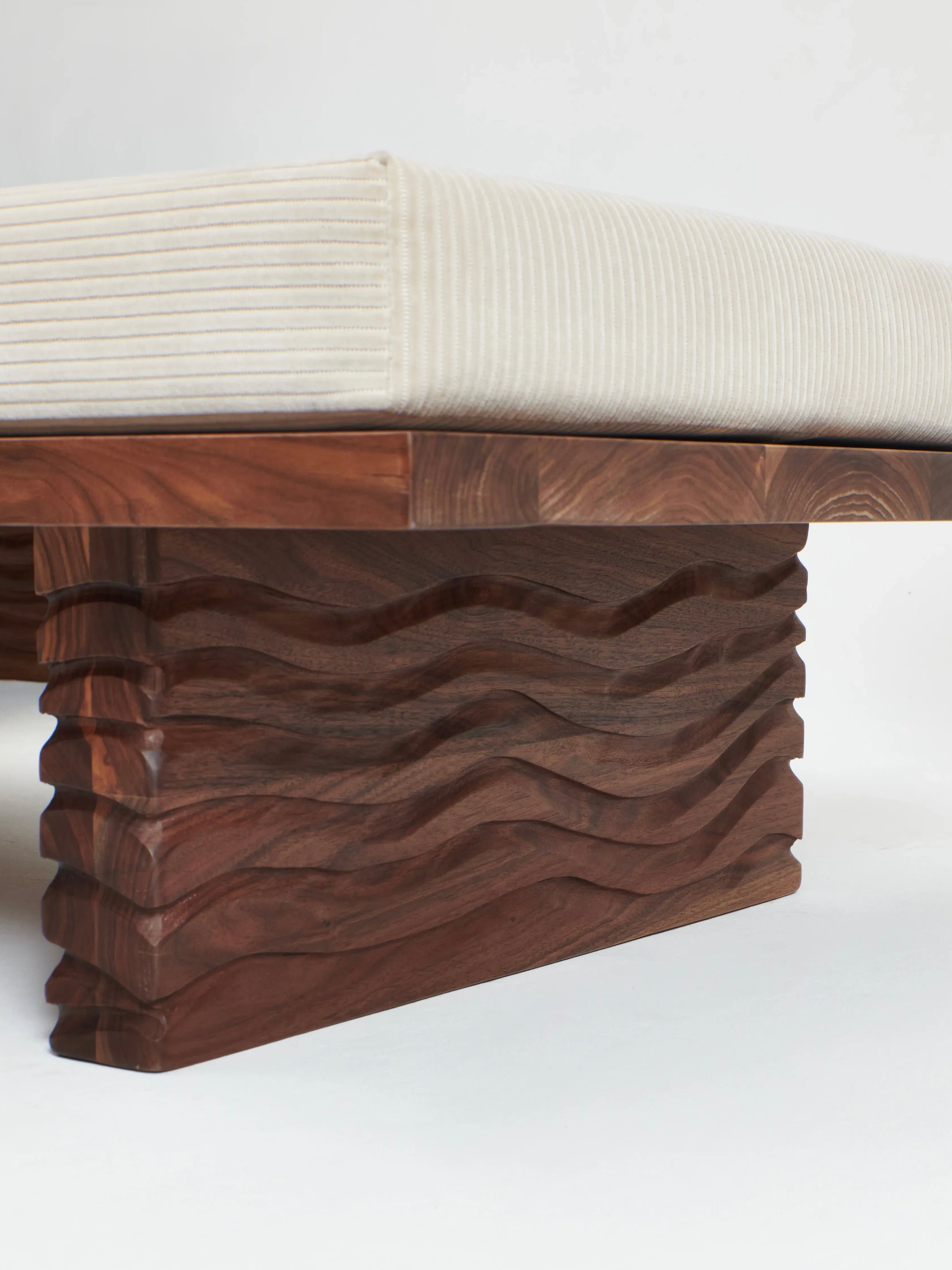 A close-up view of the Nazaré Daybed L Shape by Project 213A, handmade in Portugal, showcases a carved wavy pattern on its solid walnut legs and frame. Topped with a white, ribbed cushion, the design emphasizes craftsmanship and contrasts smooth cushioning with textured wood.