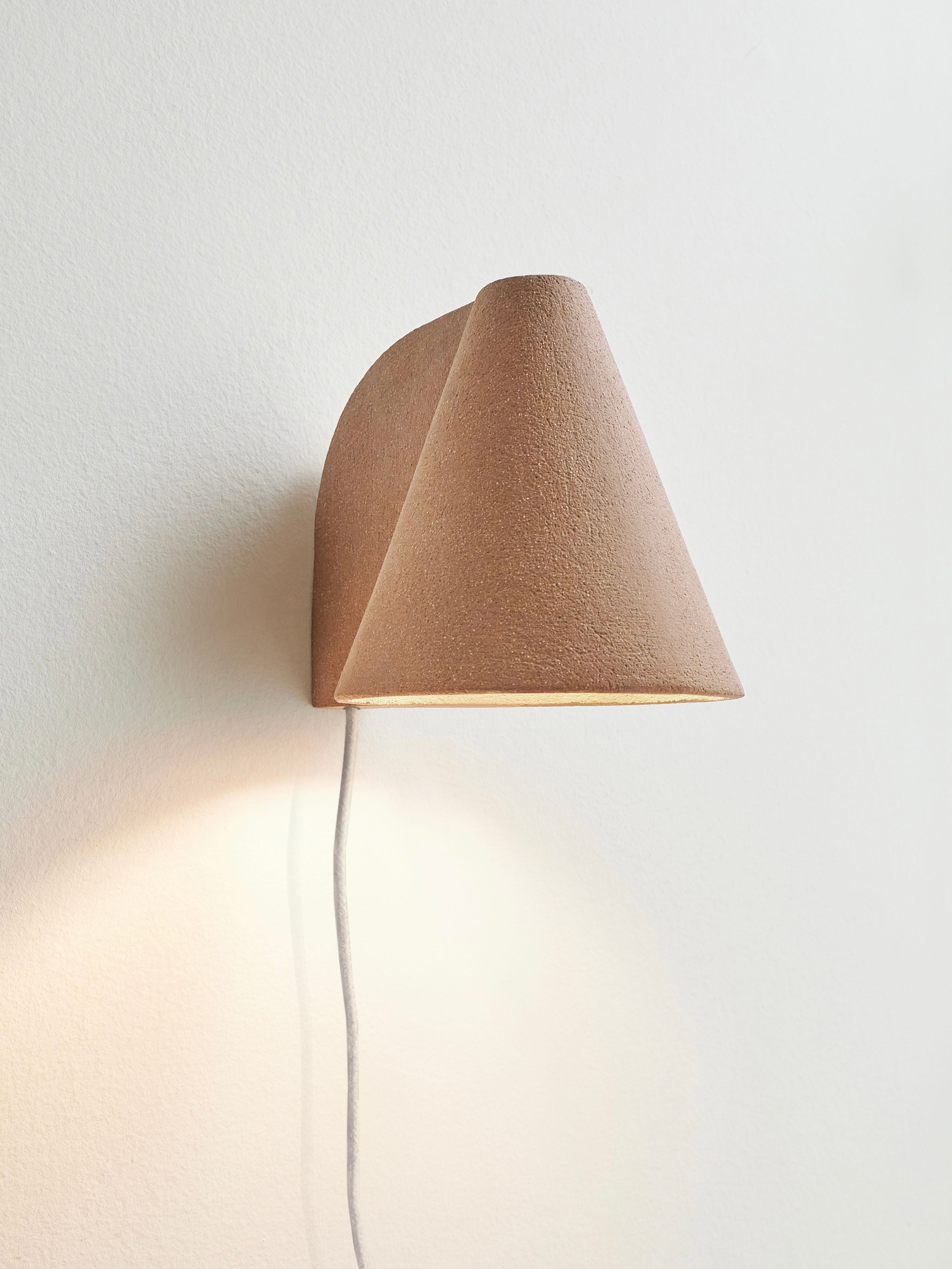 The 'Esoteric' Almond Brown Wall Light by Mariza Galani, a handcrafted cone-shaped sconce with a textured finish, is mounted on a white wall. The light emits a soft glow with a white cord hanging beneath.