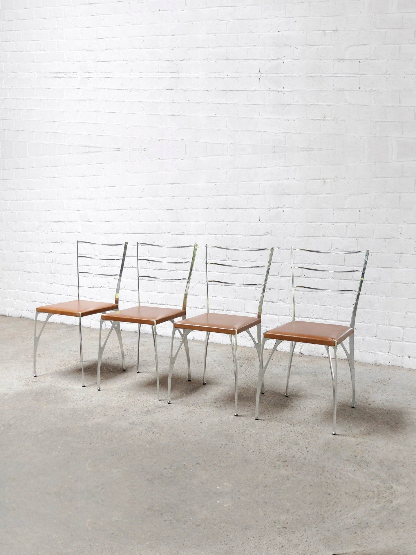 A set of four 'Organic' dining chairs by Erwan Boulloud from Introverso, featuring sculptural aluminum frames and brown seats, is arranged on a bare concrete floor against a white brick wall, enhancing the minimalist aesthetic.