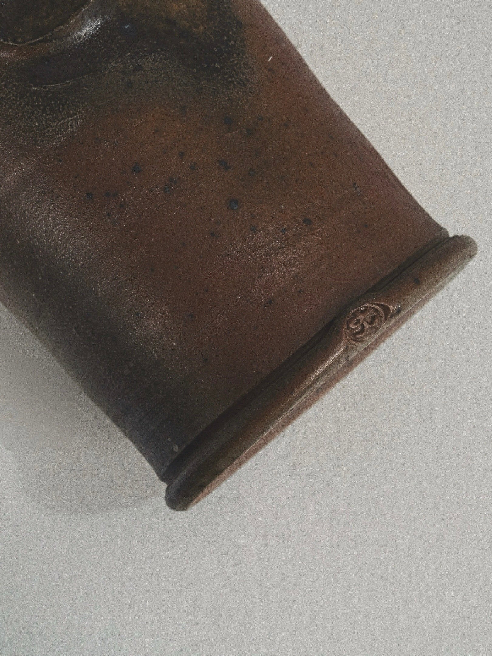 Close-up of a rustic Figuresdesign stoneware pitcher with a dark brown gradient and metallic sheen. The cylindrical end has a small stamped symbol, echoing Gustave Tiffoche's craftsmanship. The light background enhances its timeless French charm.