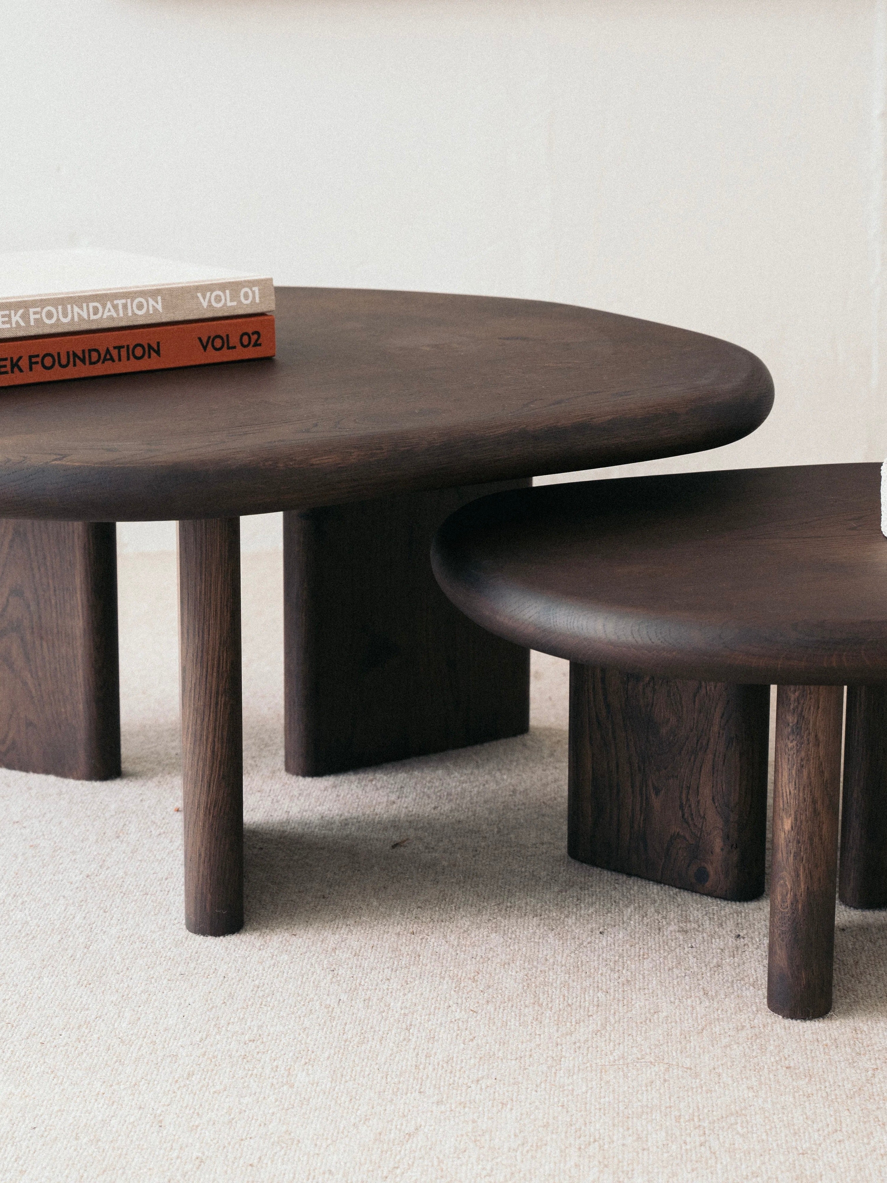 Two Double Espresso Coffee Tables by KØGE Design, crafted from solid oak with round tops and thick cylindrical legs, feature an espresso-tinted varnish finish. They are positioned on a light-colored carpet, enhancing their design curated from responsibly managed forests. A couple of books, "VOL 01" and "VOL 02," are stacked on the larger table. The minimal and light background highlights the tables' elegance.