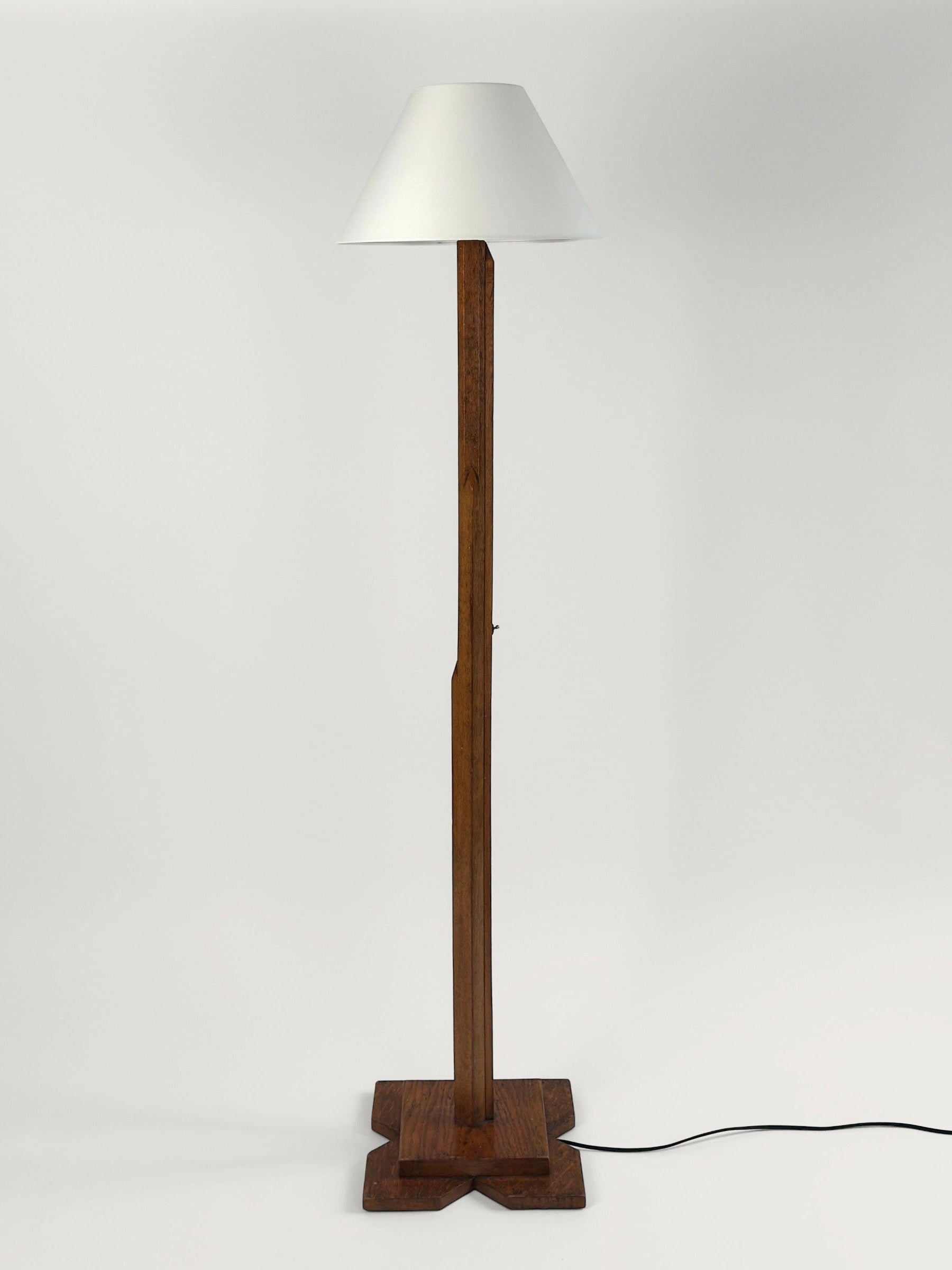 The Figuresdesign Art-Deco Floor Lamp features a white conical lampshade and stands on a cross-shaped carved wood base. A black power cord extends from the base, all set against a plain white background.