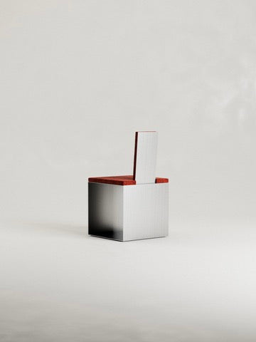 The 'Fragua' Chair by Siete Studio is a minimalist piece with a geometric design, featuring a cubic metal base and red cushioned seat and backrest against a plain light grey background.