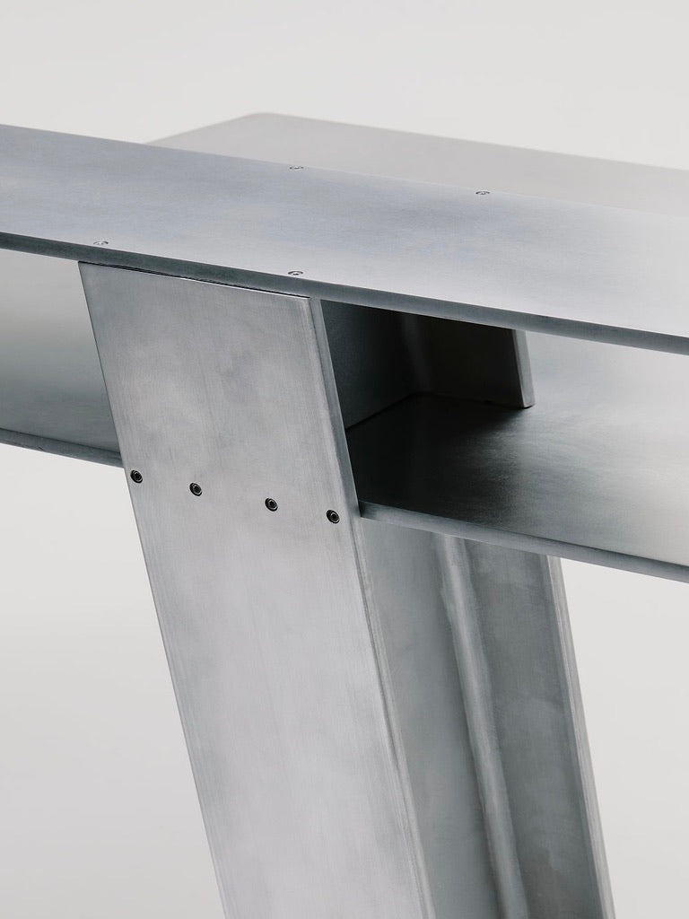 Close-up of the Beam Desk by Marquel Williams, showcasing a minimalist design with intersecting sleek aluminum sheets joined by screws. Its smooth, reflective texture and brushed finish highlight the craftsmanship.