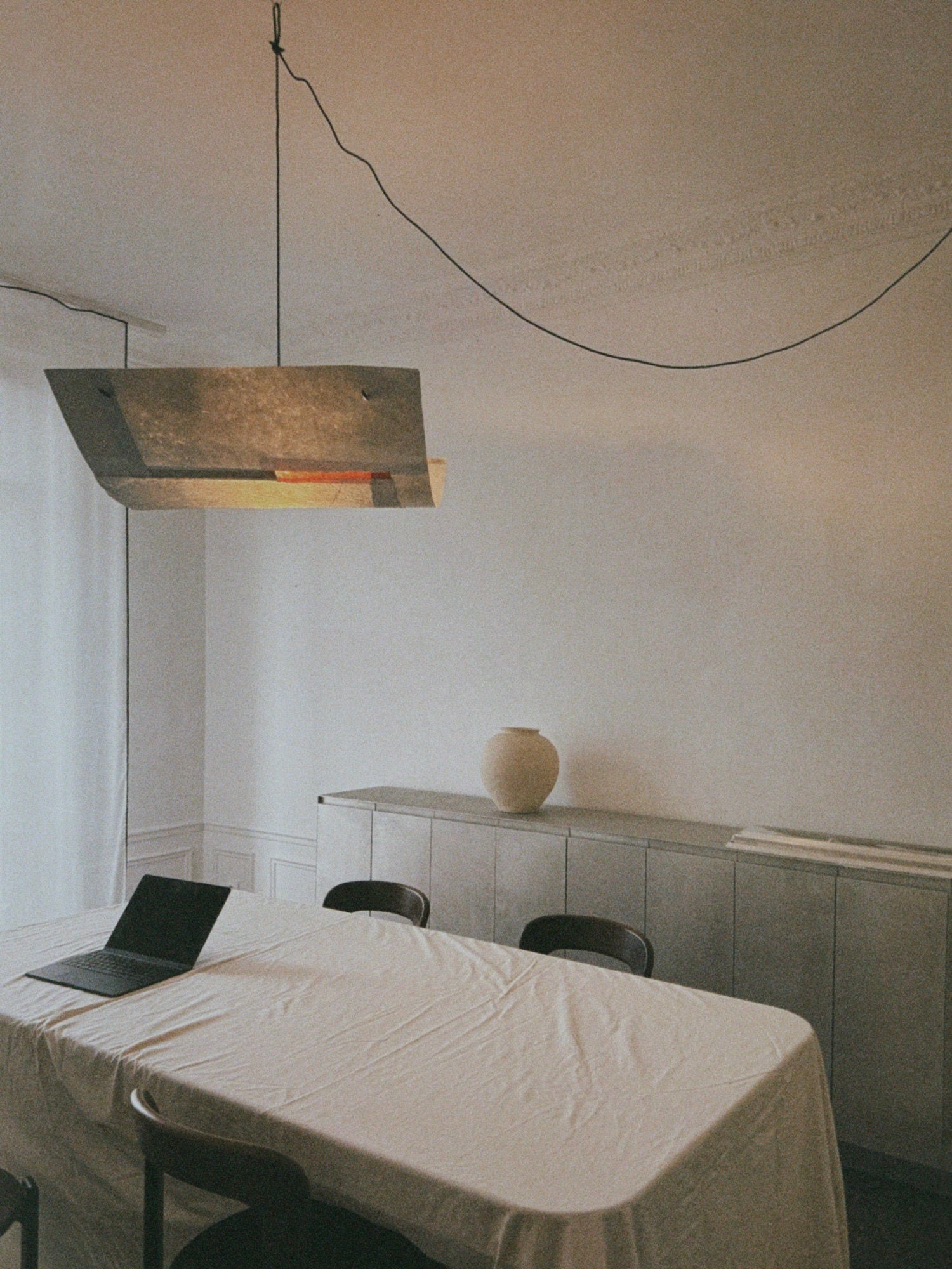 A minimalist dining room showcases a white tablecloth-covered table with a laptop and dark wooden chairs. Above, the TEGET Sail Pendant Light – Lava serves as a focal point. A vase rests on the sideboard against a plain wall, with a coiled cable visible on the ceiling.