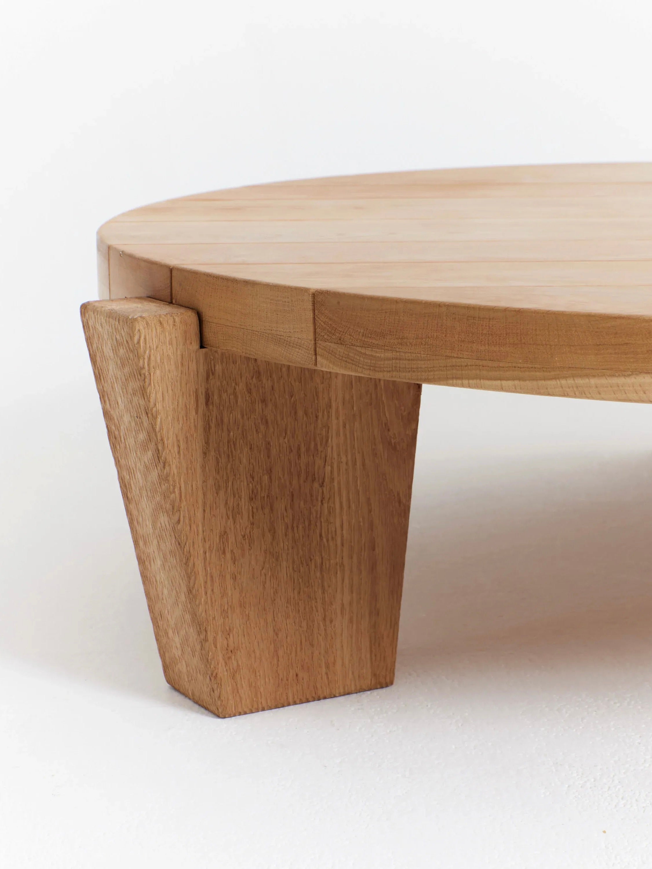 Close-up photo of the "Peniche" Coffee Table in Oak by Project 213A, handmade in Portugal from solid oak. This round, light wooden table features a slatted top and thick, angled legs that add a modern and sturdy touch to its design. The image highlights the texture and craftsmanship of the wood.