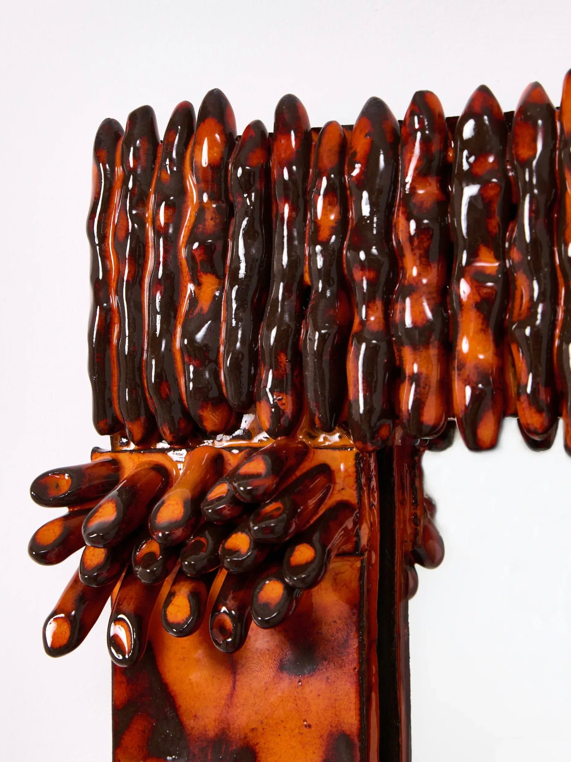 Close-up of Project 213A's 'Dedo' Ceramic Mirror, showcasing vertical elongated formations in glossy orange and dark brown tones with an organic texture, resembling finger ornaments. These handmade forms gracefully extend over a horizontal surface at the top.