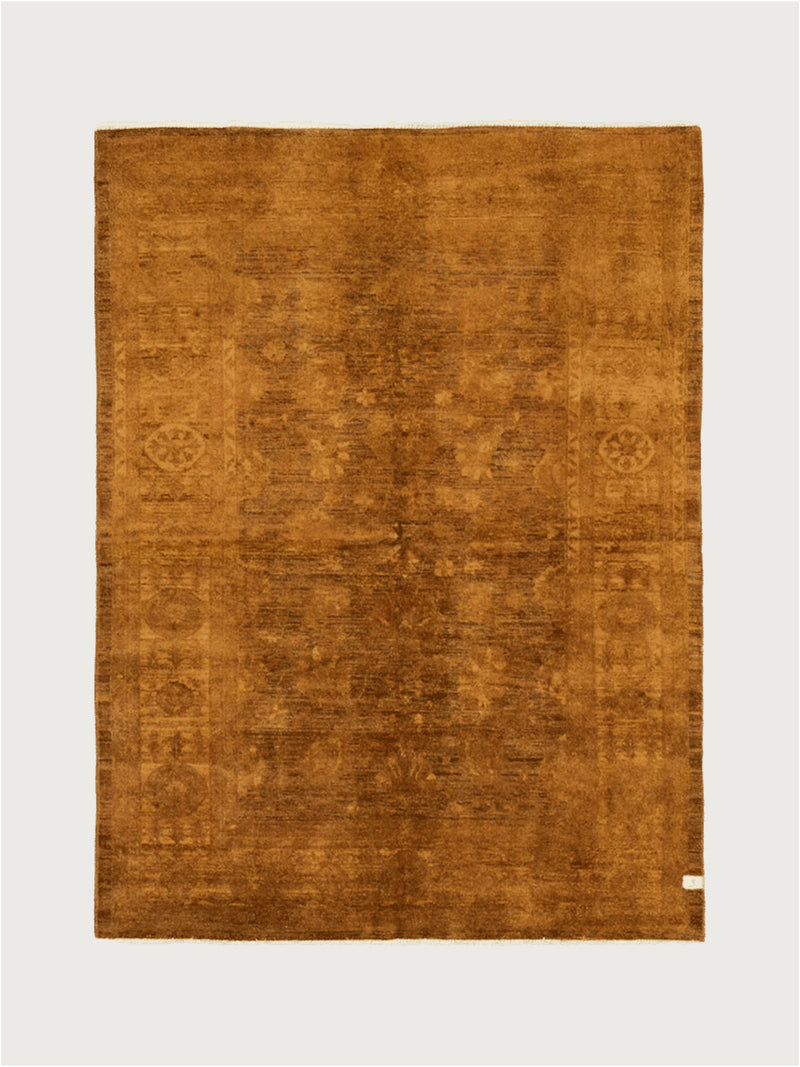 A square, hand-knotted Overdyed Rug in Safron by catalog:three, 193 x 261 cm, featuring a rich brown hue with textured wool patterns on a light neutral background and a small corner tag.