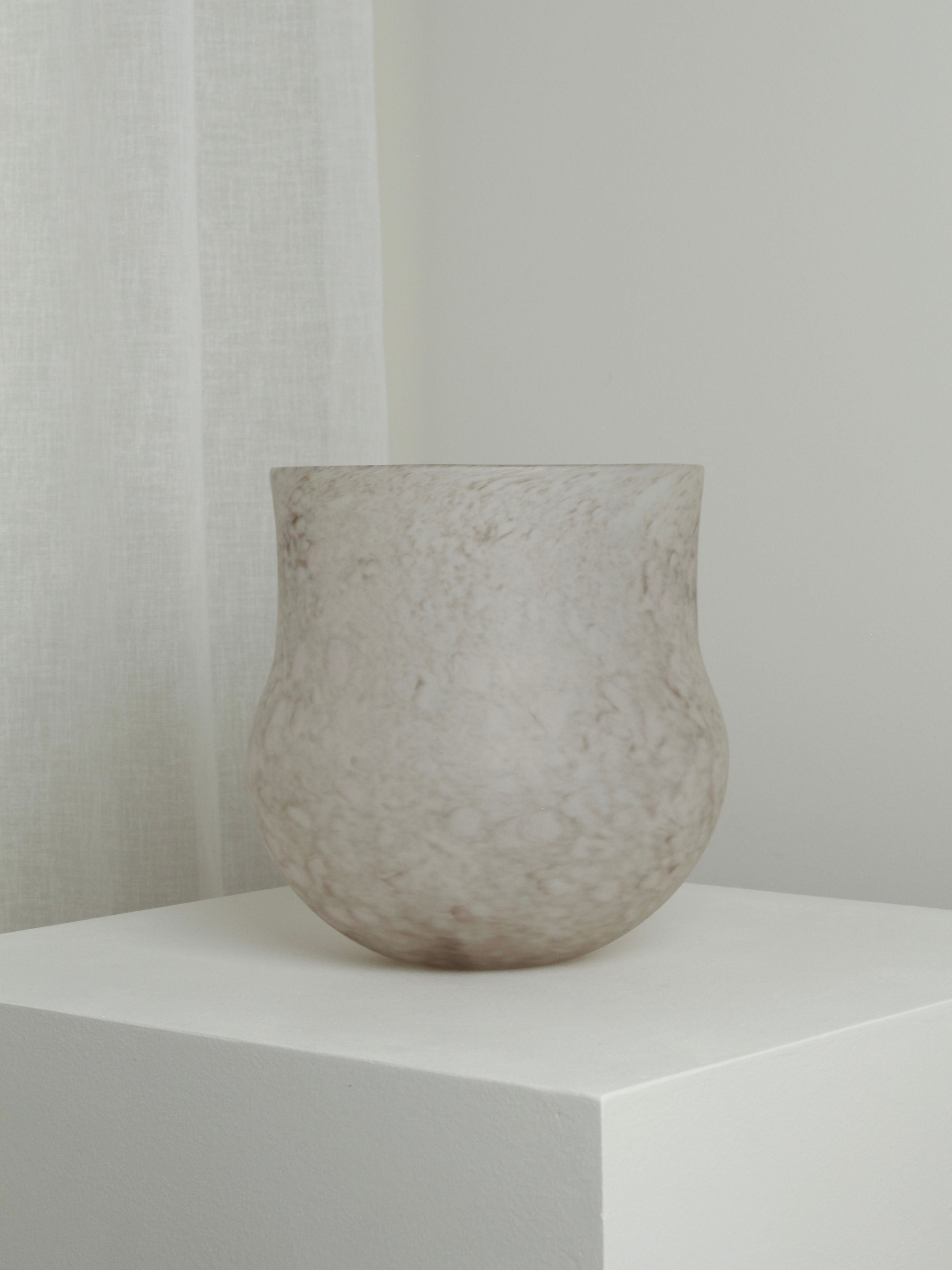 A frosted mouth-blown glass vase with a bulbous base and a slightly narrowed, flat rim sits on a white pedestal. The background features a soft, sheer white curtain and a light gray wall. The scene is minimalistic and calm, showcasing the Verre d'Onge XLarge Vase in Feathered Taupe beautifully.