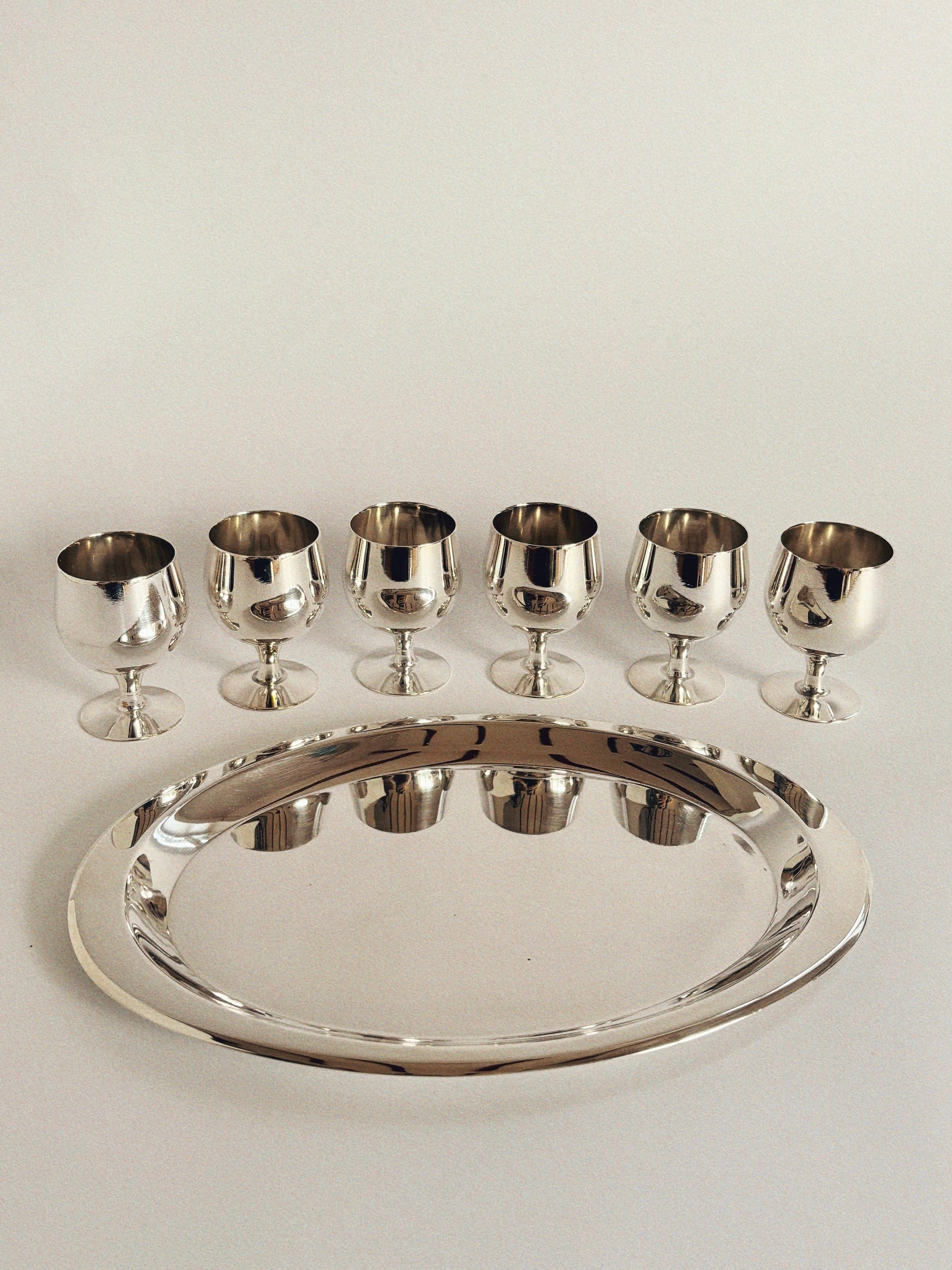 The Silver Plated 70's Liquor Server by Huerto is beautifully displayed with six silver-plated liquor glasses neatly aligned on an oval matching tray against a plain white background, their metallic surfaces gleaming as they catch the light.