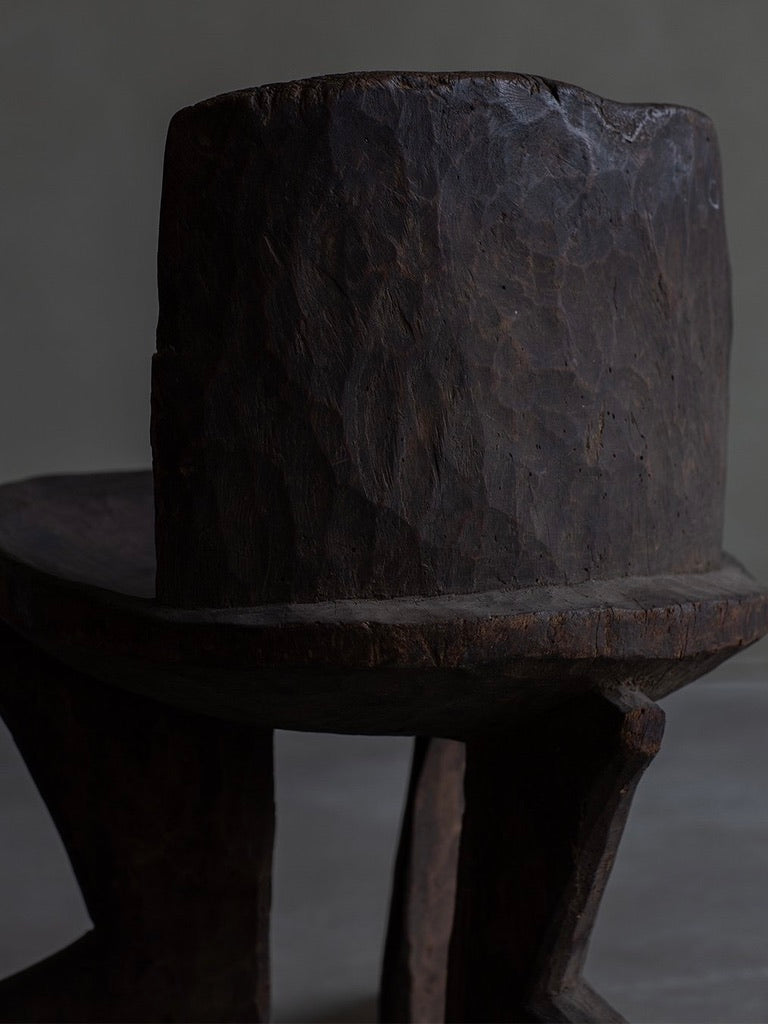 The African Stool by Bicci de' Medici features a rustic design with a thick carved backrest and sturdy legs. Its dark wooden surface reveals visible textures and natural imperfections, showcasing the skilled craftsmanship of African artisans against a muted background.