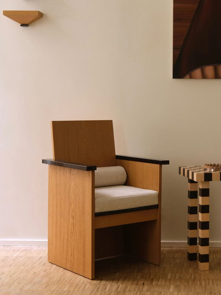 A modern minimalist room features the "Le Parfait" armchair by HAUVETTE & MADANI, showcasing lacquered armrests and a beige cushion. A small striped side table complements the setting along with a wall-mounted shelf on a light-colored wall, while the brushed oak floor adds warmth to the decor.