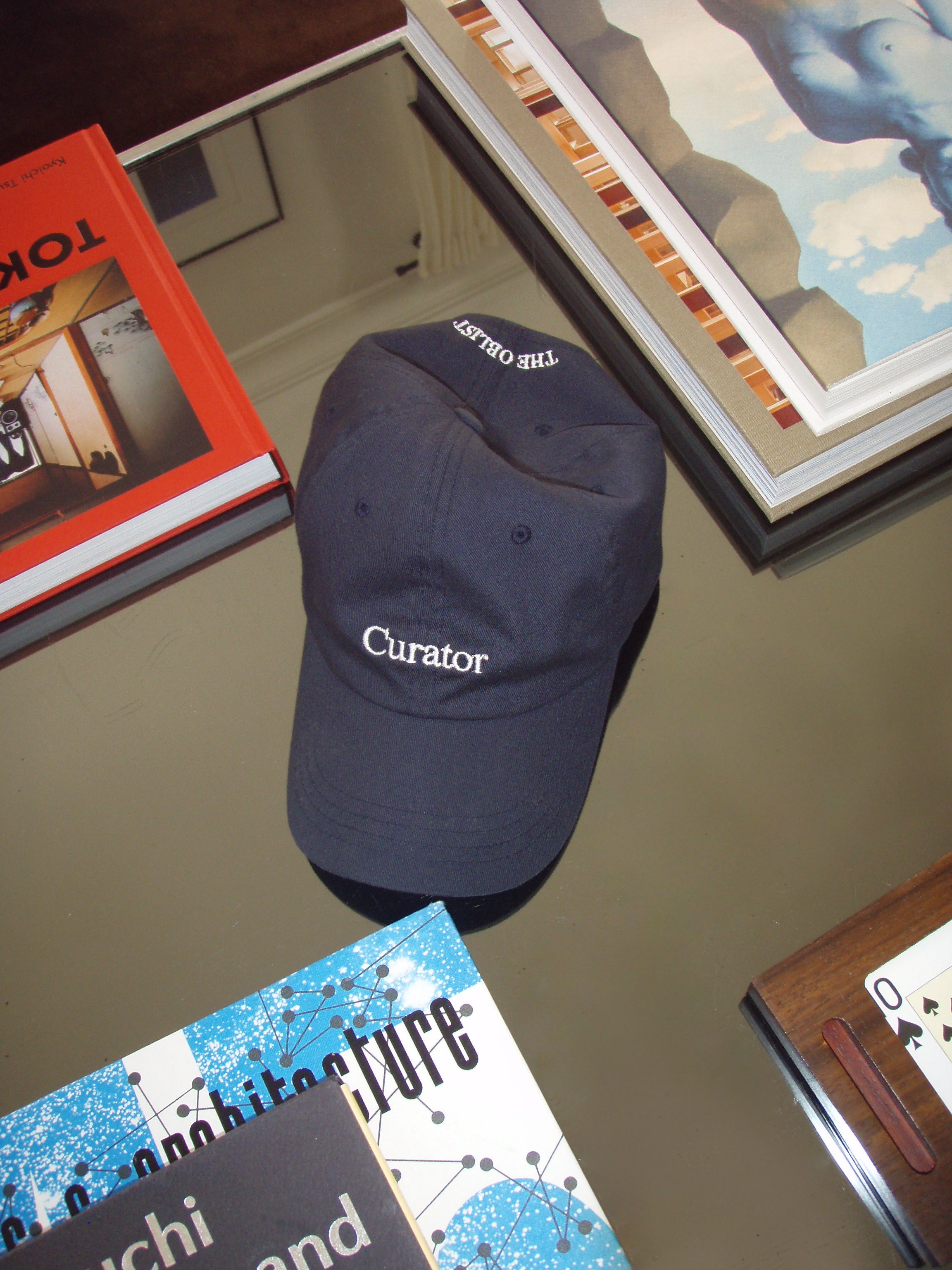 A black cap, labeled "The Curated Cap" from The Oblist, featuring "Curator" on the front and "THE OUTLIER" on the back, sits elegantly on a reflective glass table. Its unisex design effortlessly complements nearby art books and a partially revealed painting. This limited edition piece is crafted with breathable fabric to ensure comfort.