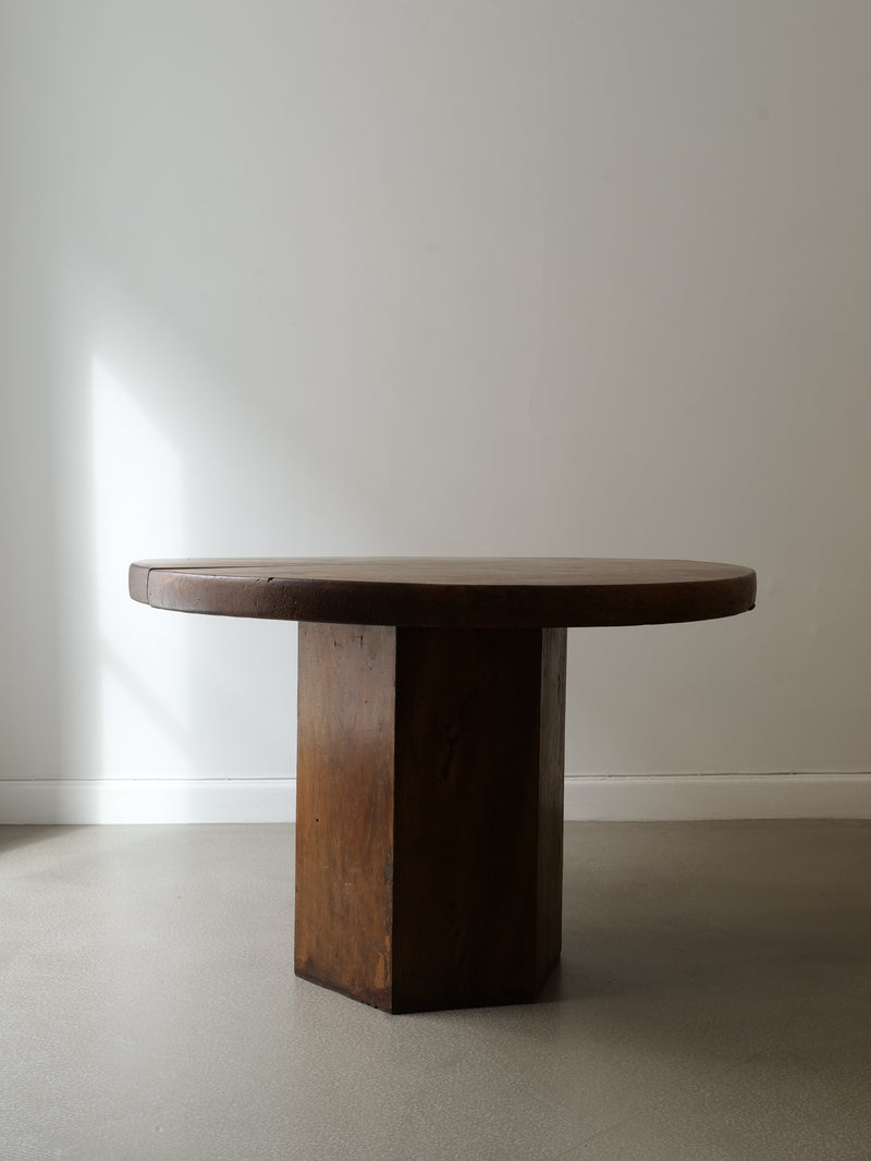 The Collection apart Midcentury Hexagonal Wood Dining Table, featuring a thick top and sturdy rectangular pedestal base, stands in a minimalistic room with light gray floors and white walls as soft light filters in from the left.