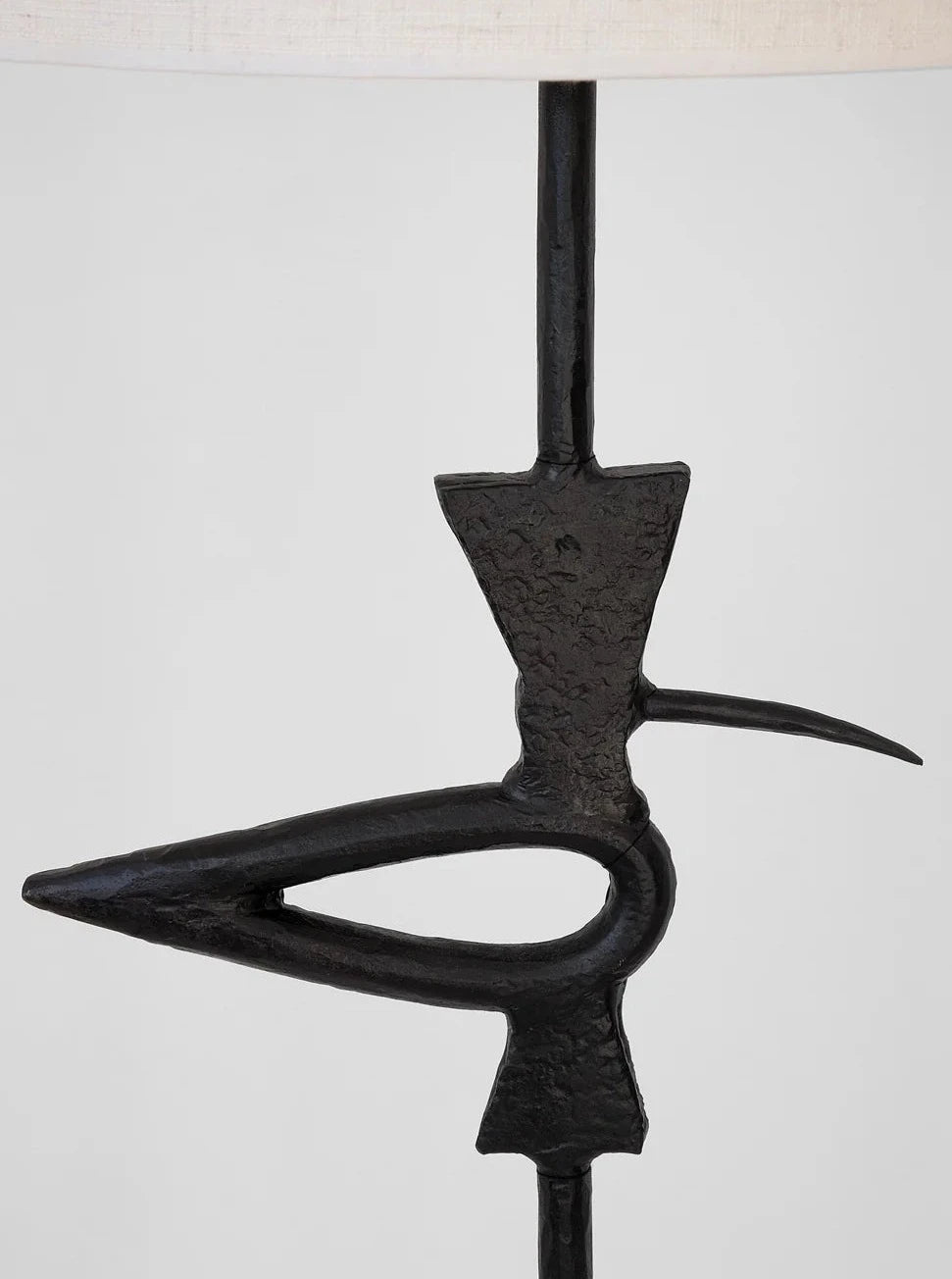 A close-up photo of an abstract, black metal sculpture resembling a bird with a pointed beak and an elongated, curved body. The "Brejos" Floor Lamp by Barracuda Interiors features this sculpture attached to a vertical rod, set against a plain light gray background.