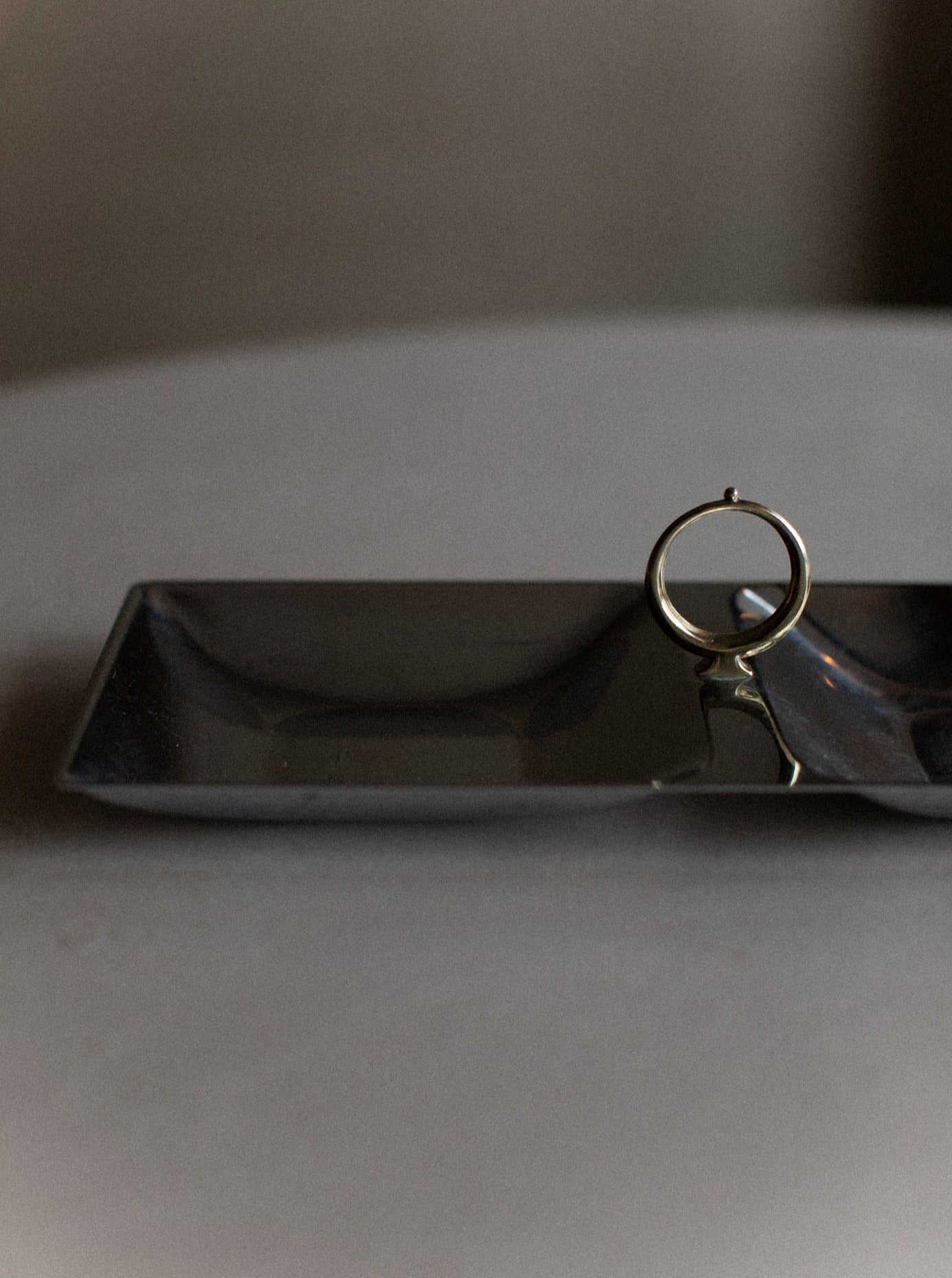 A minimalist scene featuring a thin gold ring standing upright on one edge of a sleek, matte black Out For Lunch Stainless Tray exudes modern elegance. The softly blurred background enhances the contemporary style, giving the composition a serene and elegant feel.