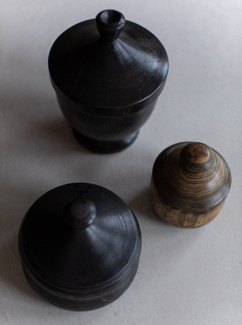 A top-down view of three wooden containers with lids, placed on a light surface. The **Set of Three Boxes** by **Out For Lunch** exudes rustic elegance with their simple, handcrafted appearance, each showcasing unique textures and natural wood grain. The two larger ones are darker, while the smallest one features a lighter finish.