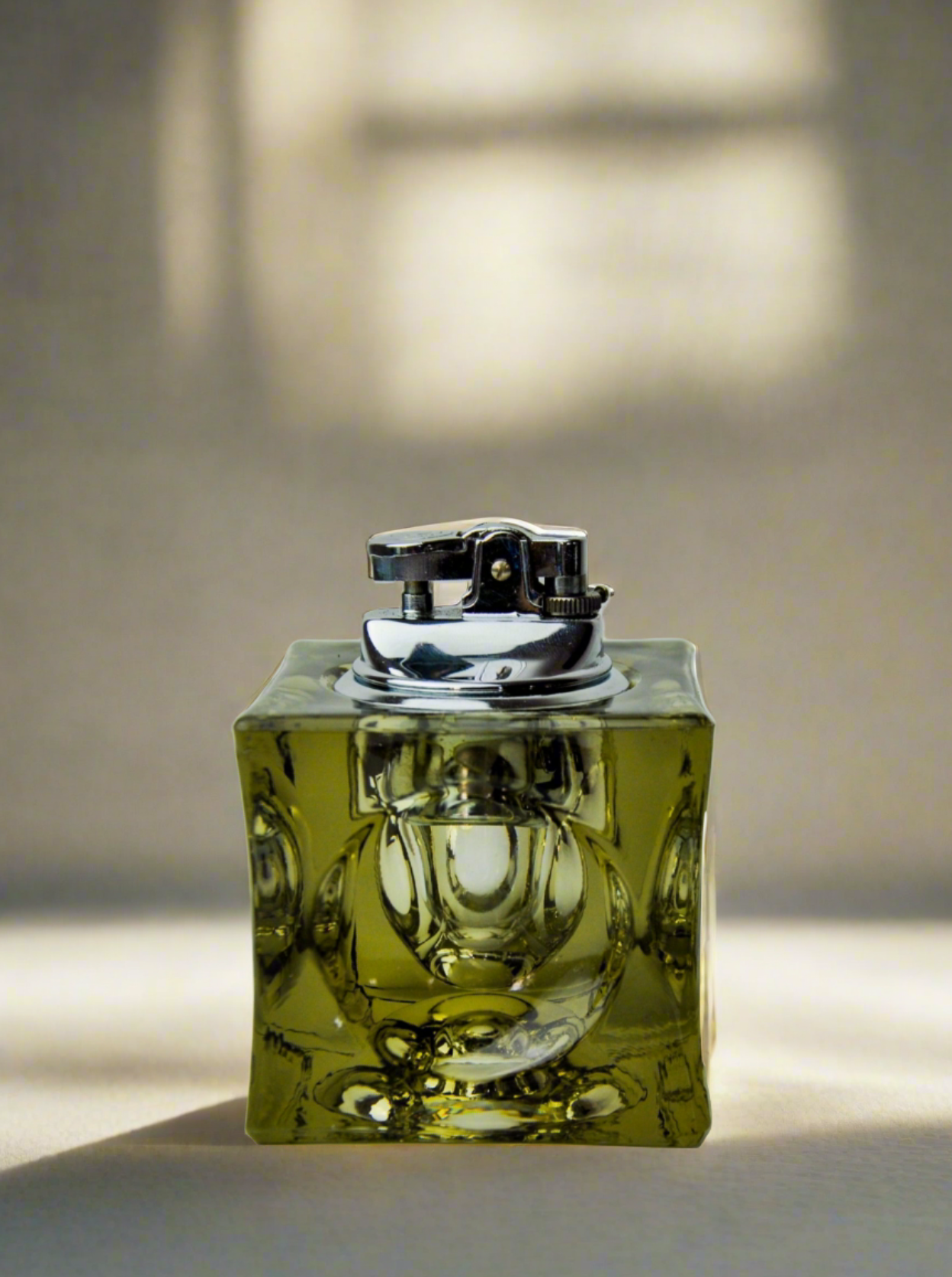 A vintage square glass lighter, known as the Ice Cube Lighter by Antonio Imperatore (1970), containing a yellow-green liquid, stands upright against a plain white background. Created by Venetian master glassmakers from Malamar Studio, the lighter features a silver top with a flint wheel and an intricate pattern on the glass body that exemplifies Antonio Imperatore's craftsmanship.